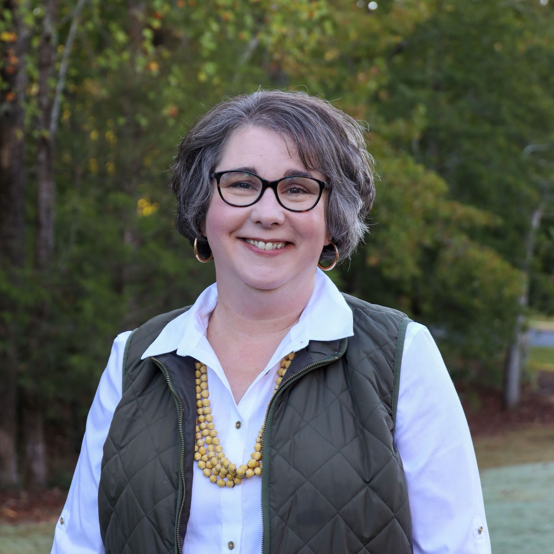 Smiling person with glasses in a white shirt and quilted vest standing outdoors on a leafy path