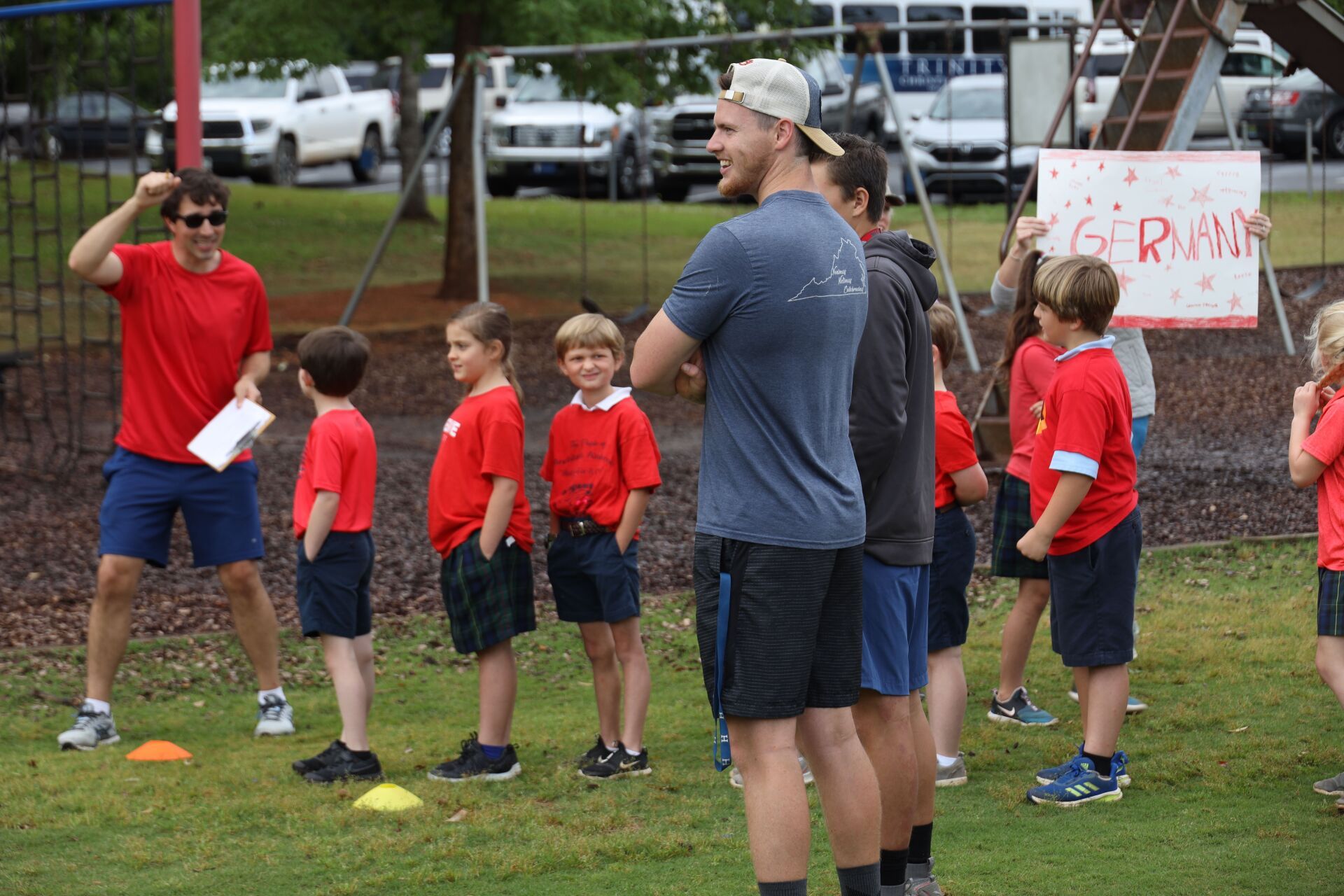 Children in red shirts stand on a grassy field while adults coach them near sports cones and tents.