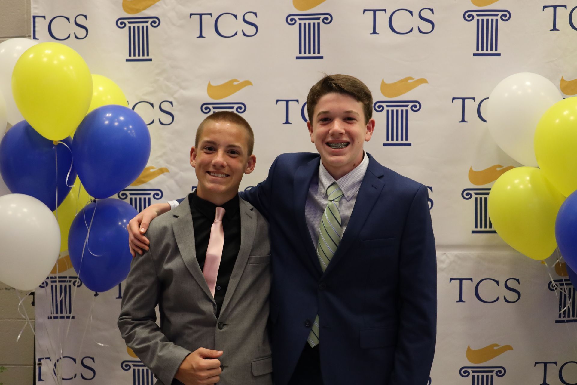Two people in suits pose in front of a TCS backdrop with blue, yellow, and white balloons
