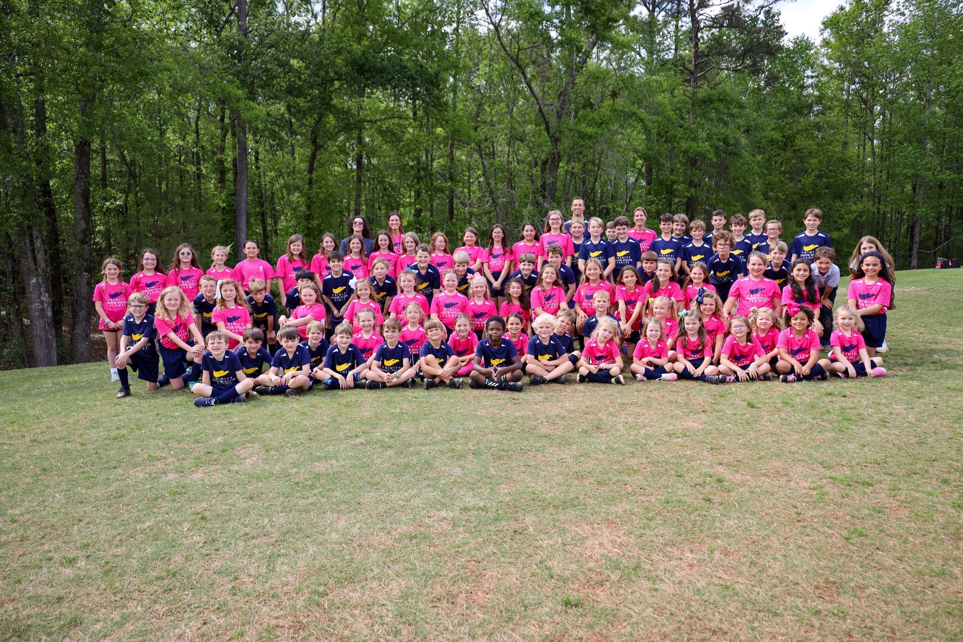 Large group in pink and dark shirts posing outdoors on grass in front of trees