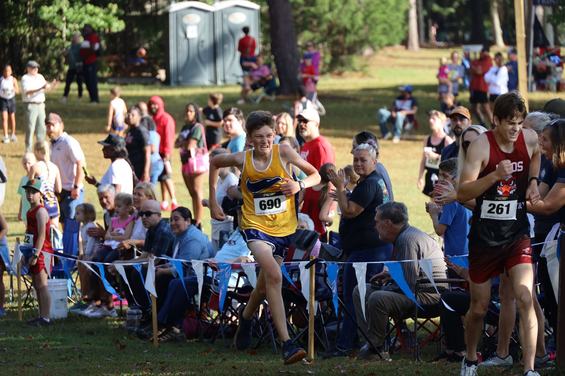 Runners race along a grassy course beside spectators seated behind a rope barrier.