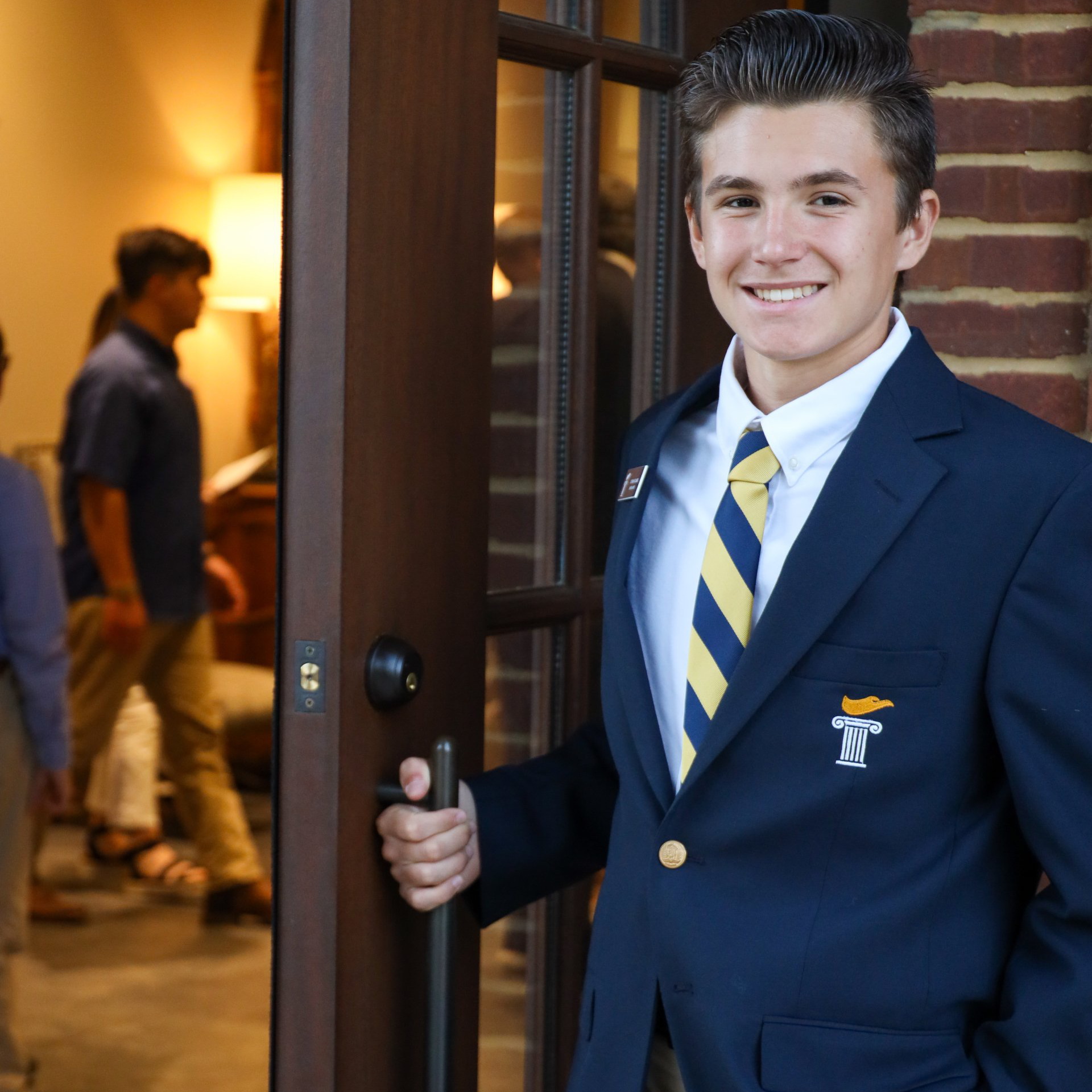 Young man in navy blazer and striped tie opens a doorway, smiling, with a brick building behind him.