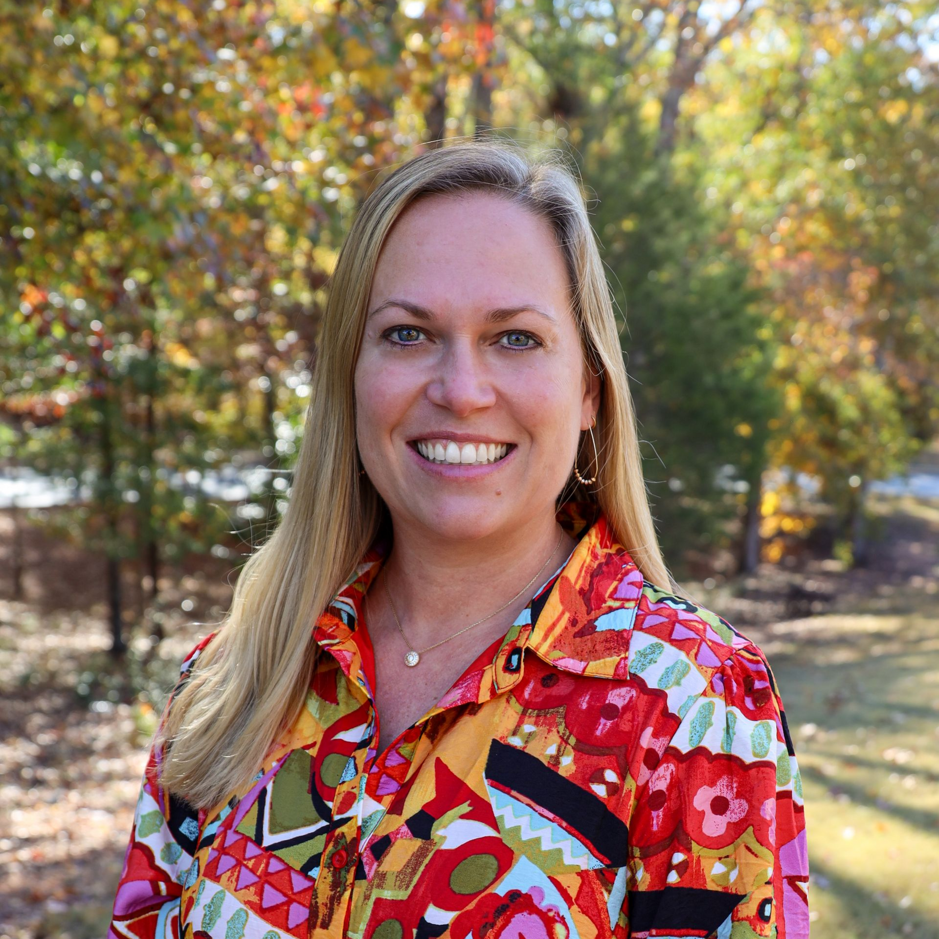 Smiling woman in a colorful shirt standing outdoors with autumn trees in the background