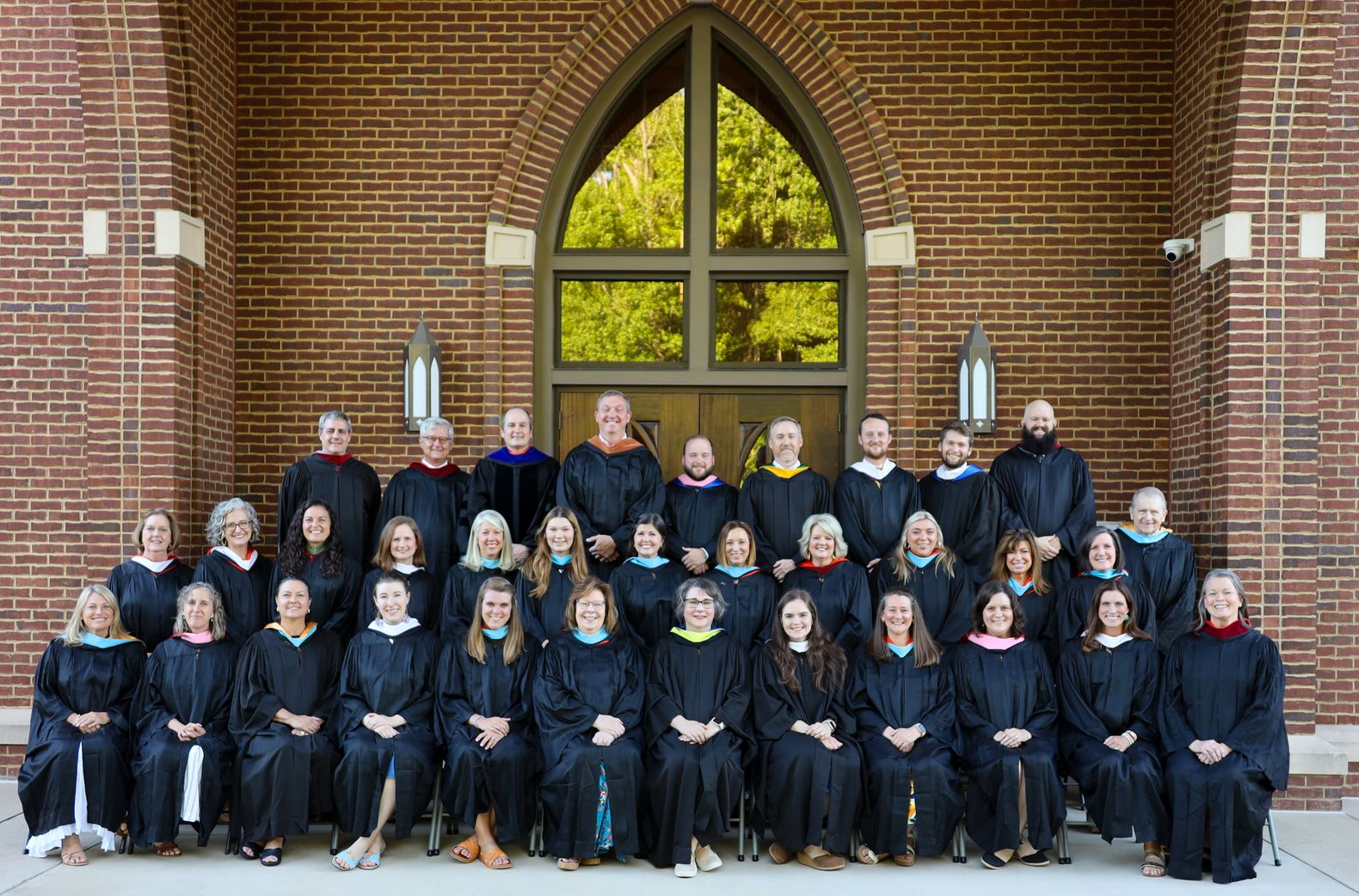 Group portrait of people in dark robes seated and standing in front of a brick building archway