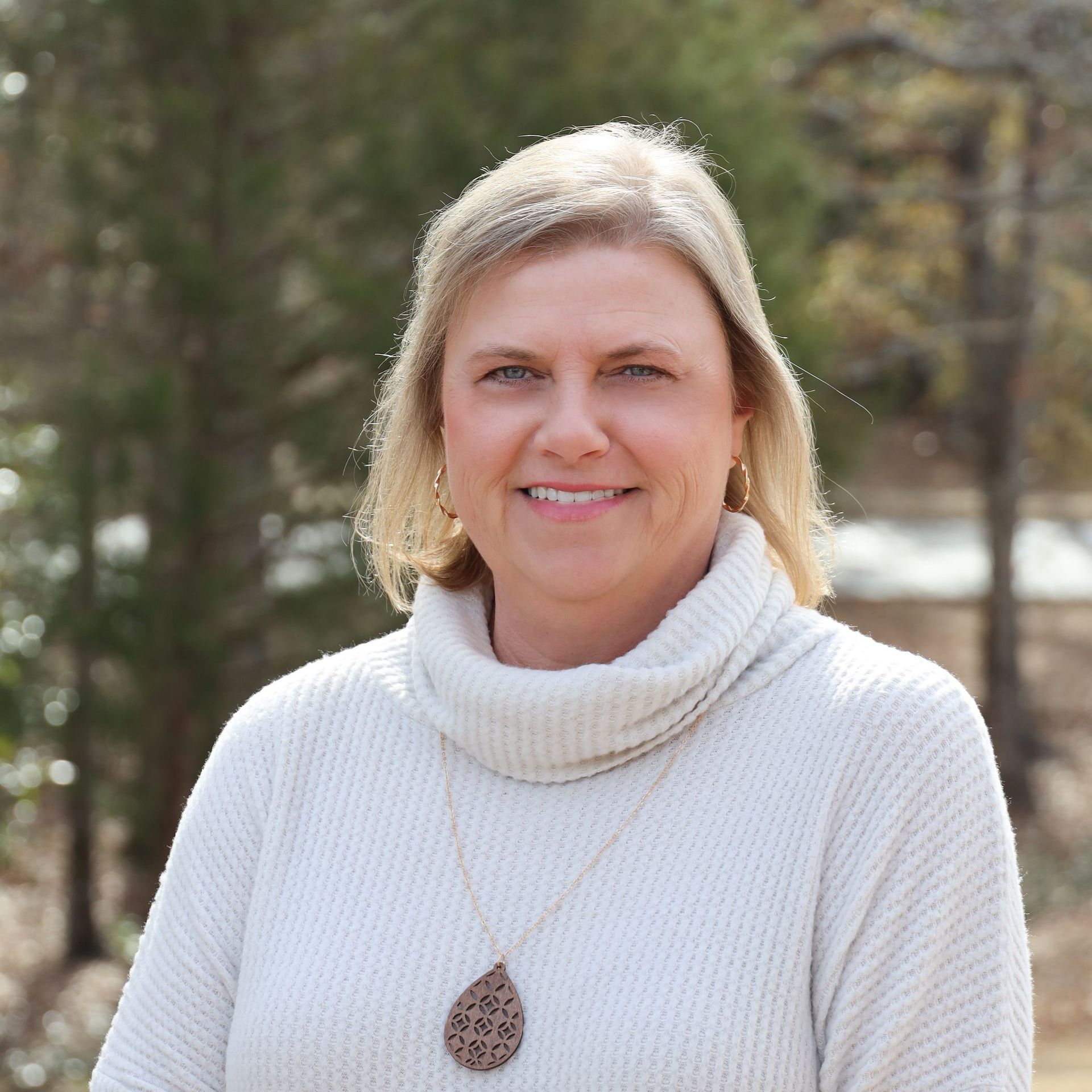 Smiling woman in a white sweater and necklace standing outdoors with trees in the background