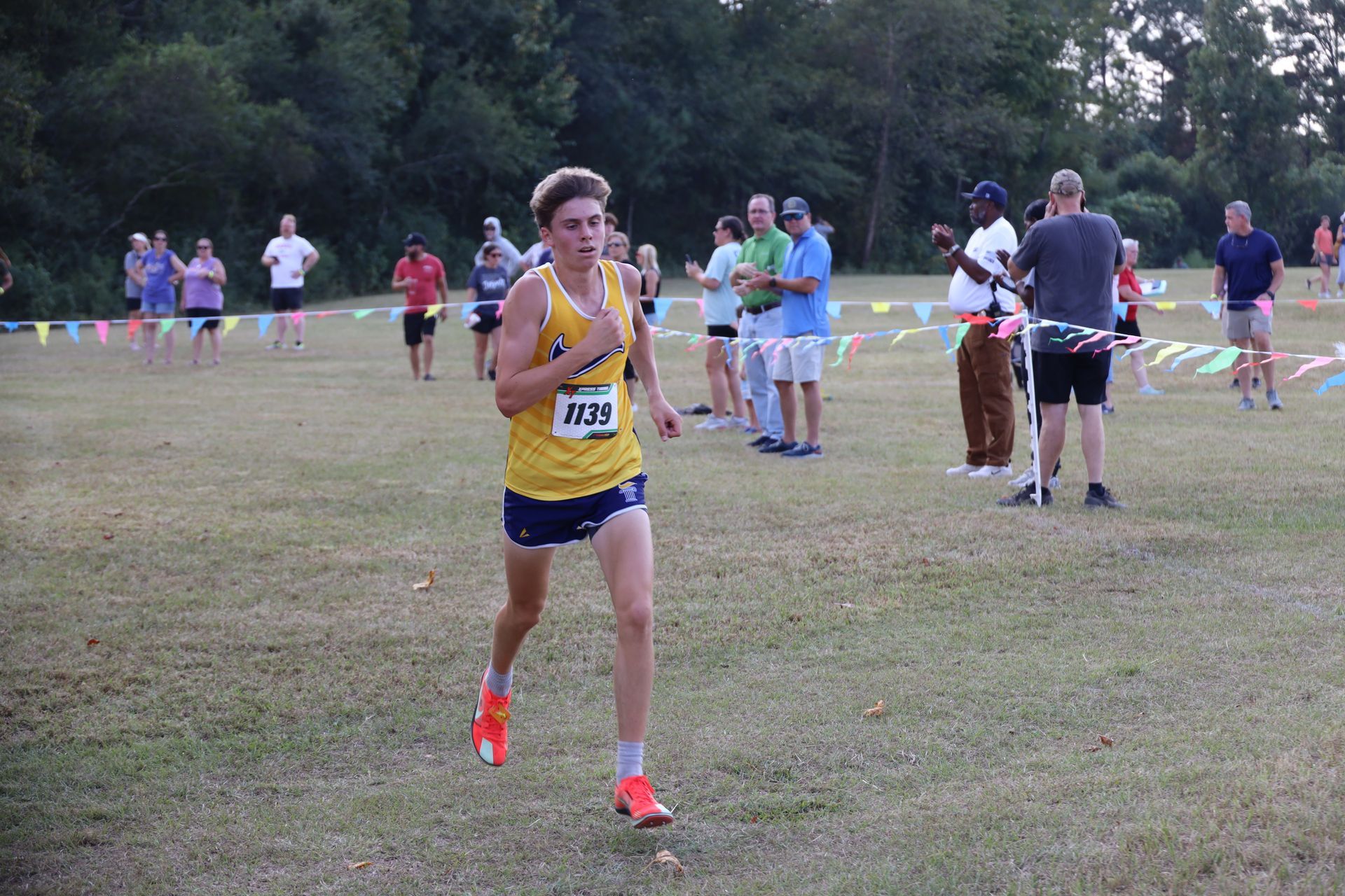 Runner in blue-and-yellow uniform races past spectators at a grassy cross-country finish area.