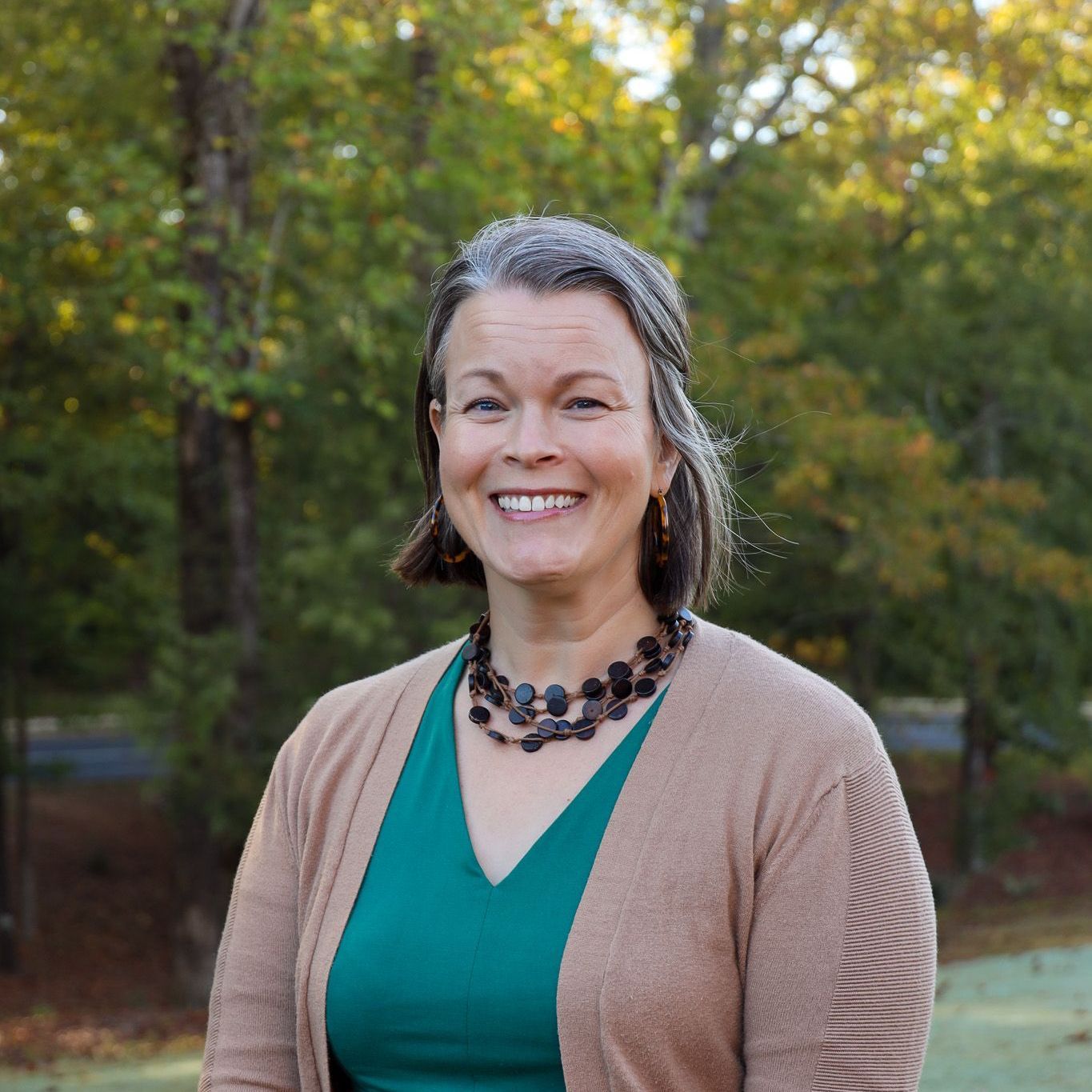 Smiling person in teal top and beige cardigan standing outdoors with trees in the background