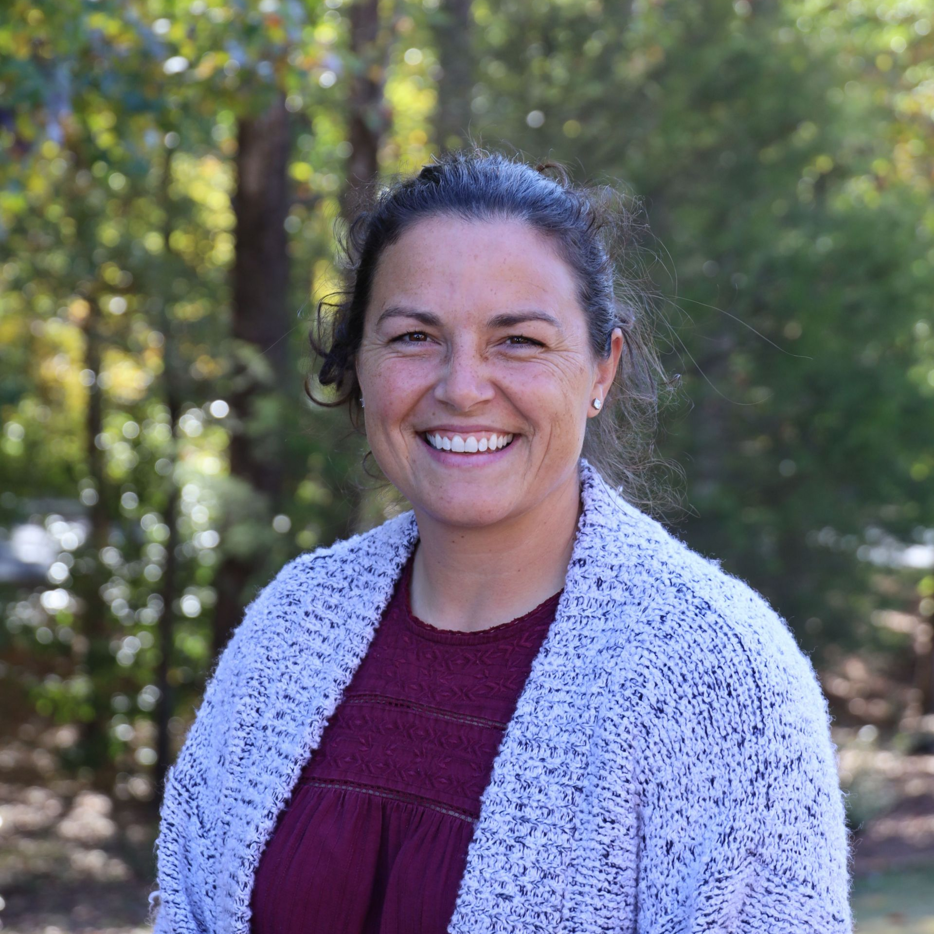 Smiling woman in a maroon top and gray cardigan standing outdoors with trees in the background