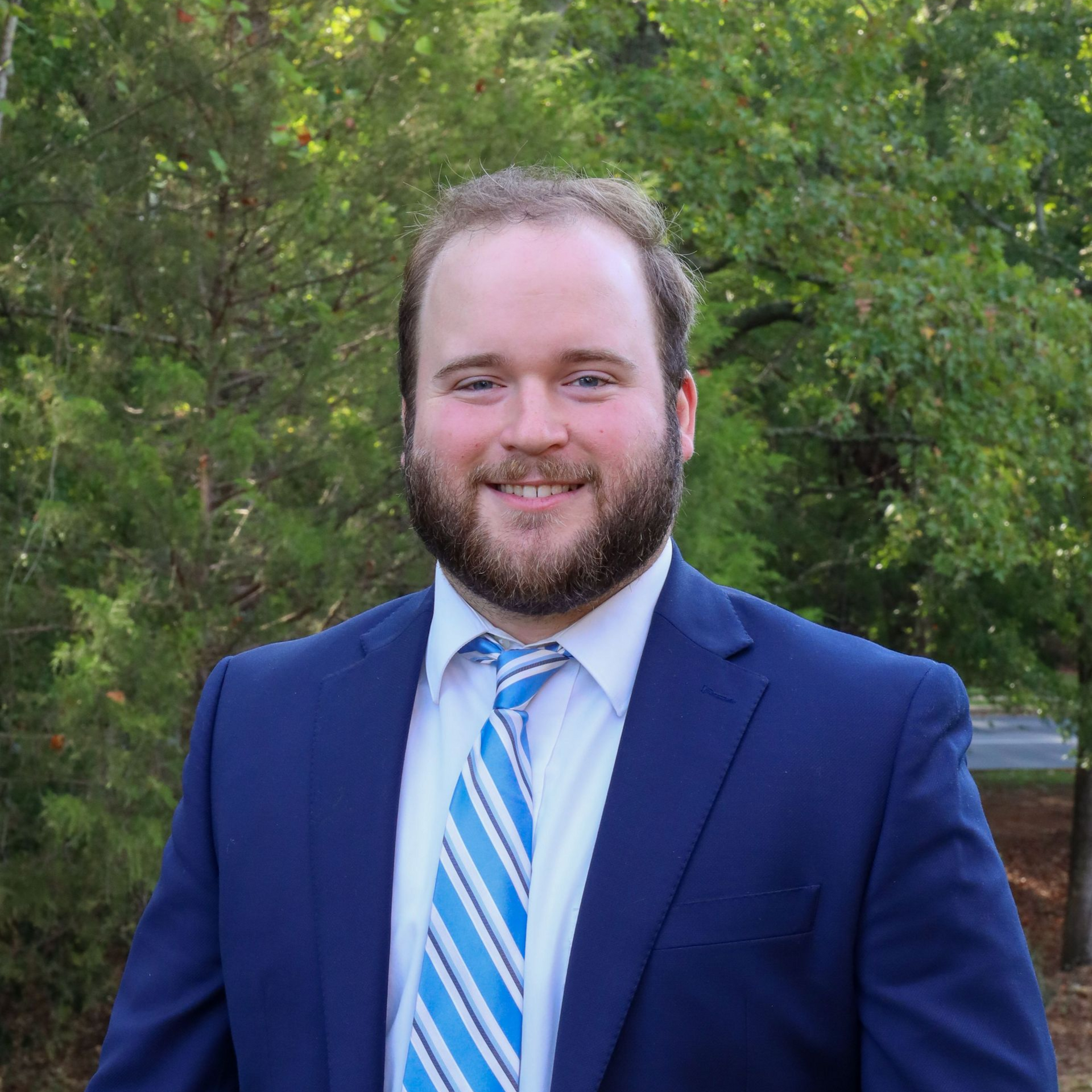 Smiling man in a blue suit and striped tie standing outdoors in front of green trees