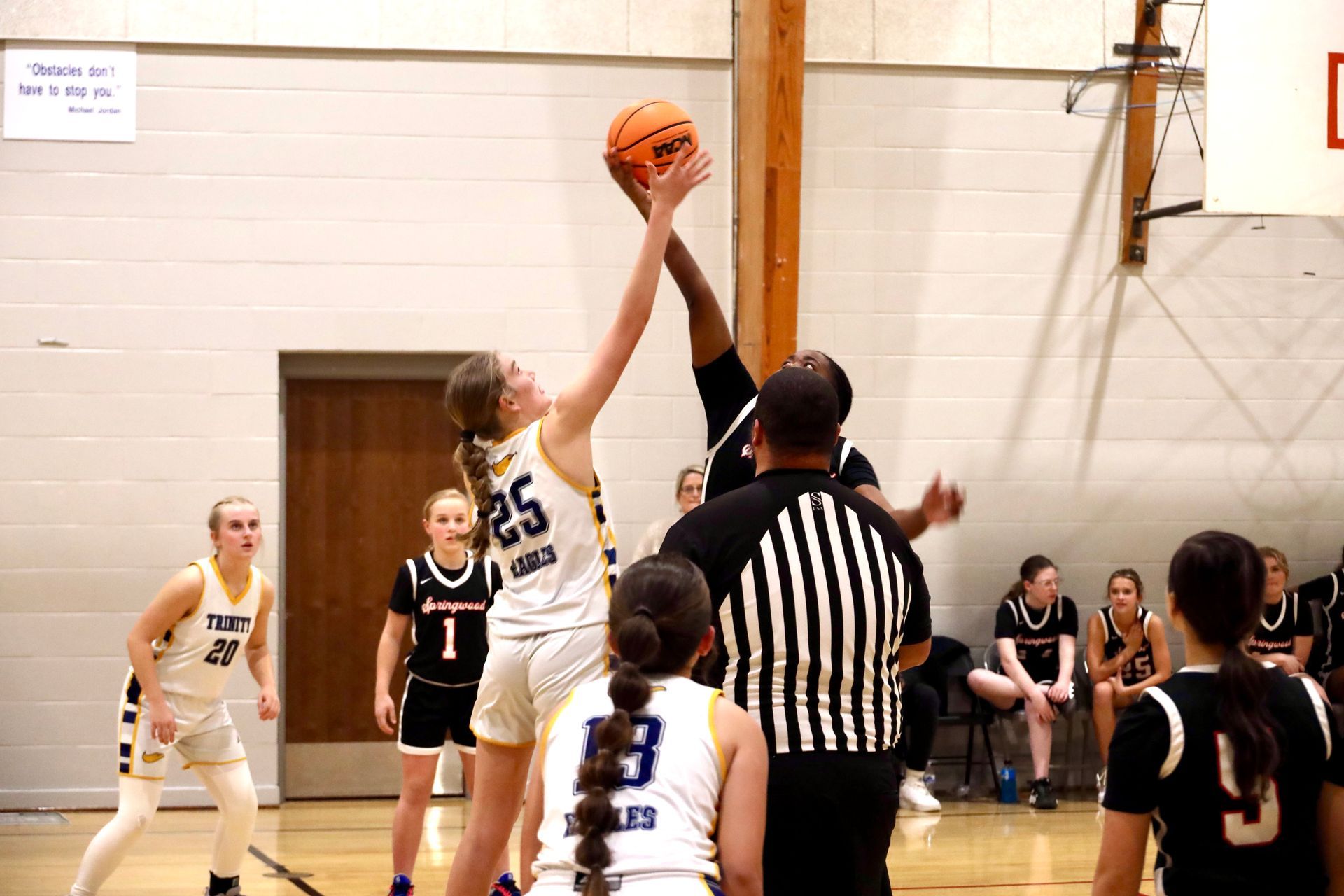 Basketball tip-off in a gym, referee between two players reaching for the ball.