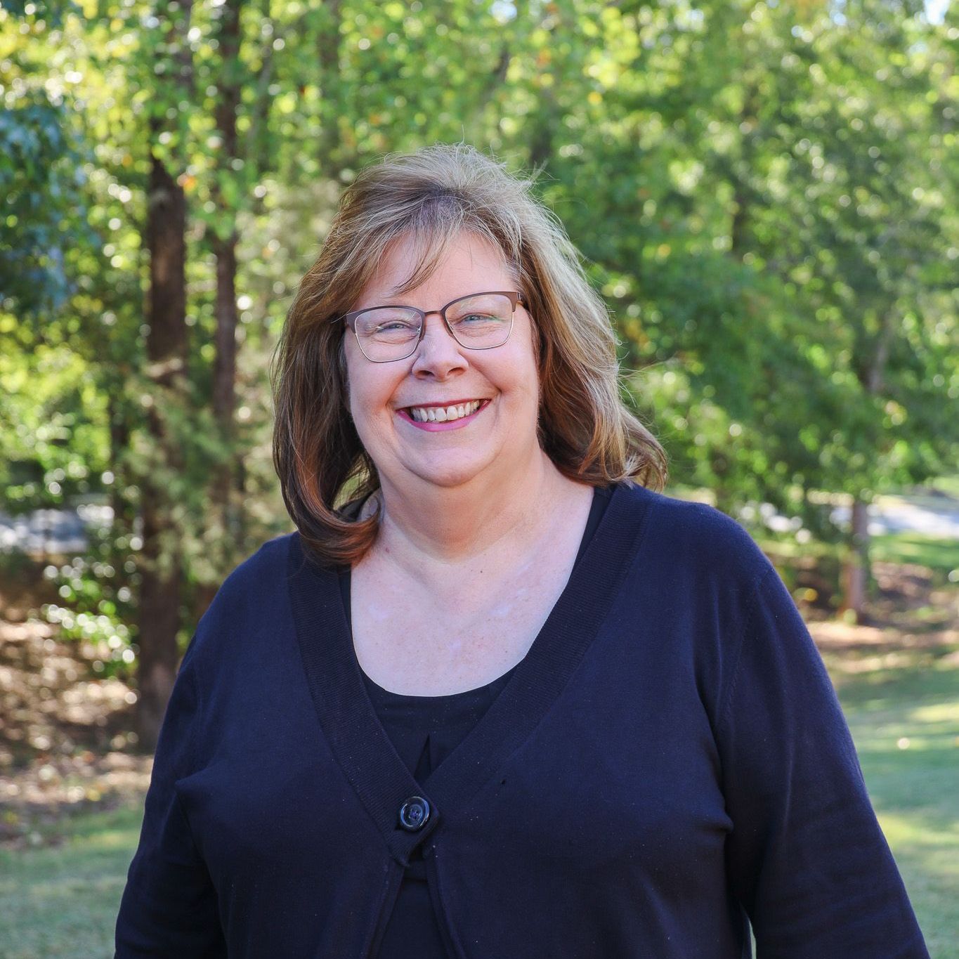 Smiling woman in glasses standing outdoors among green trees, wearing a dark top.