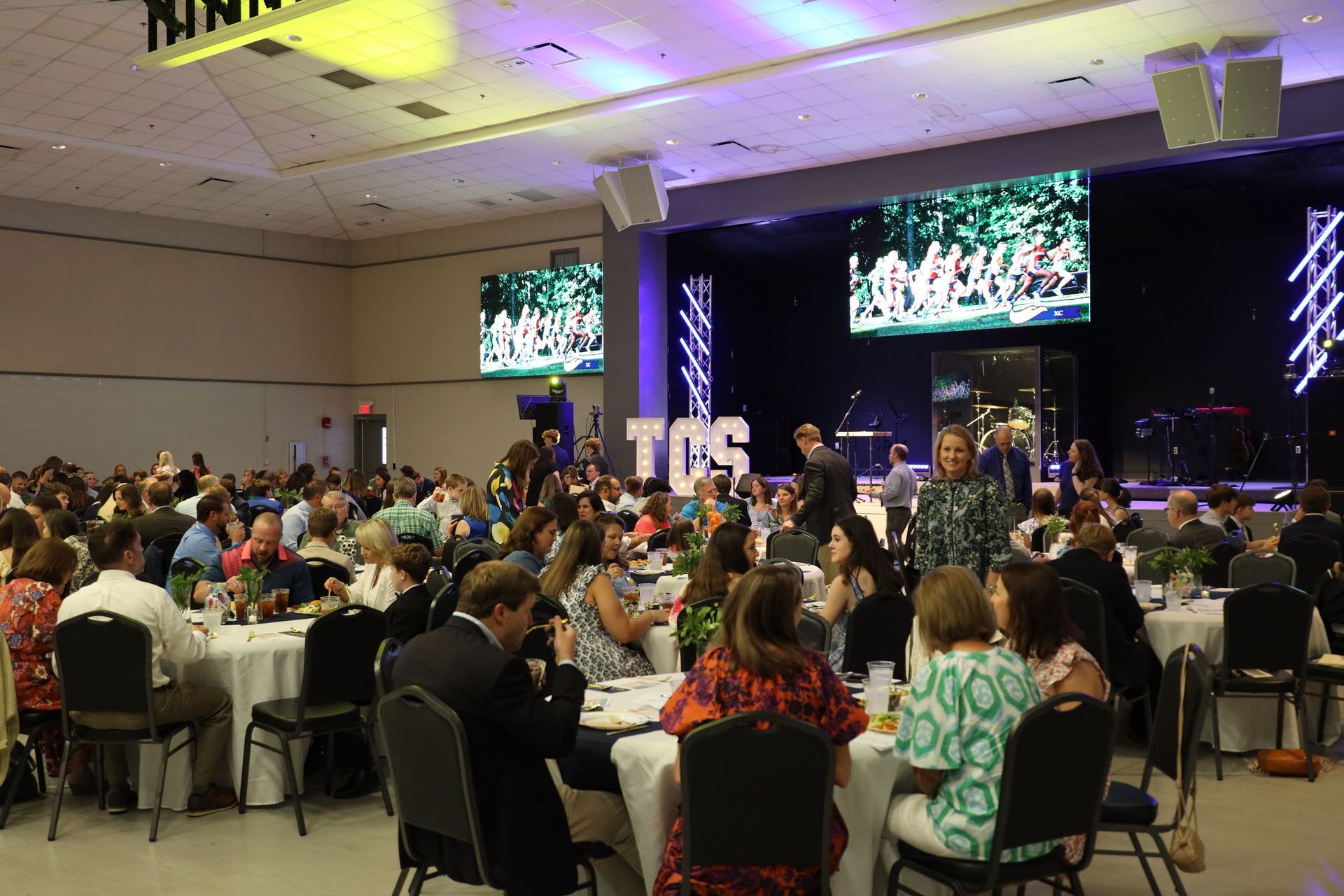 Conference banquet hall with seated crowd facing a stage and large screens displaying a live performance.