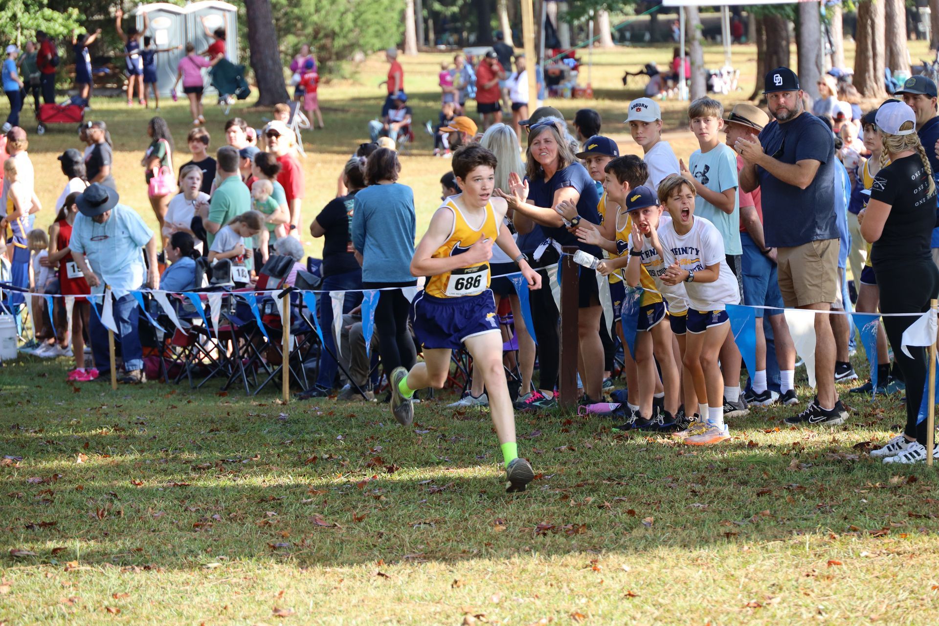 Runner in blue-and-yellow uniform races past spectators at a grassy cross-country finish area.