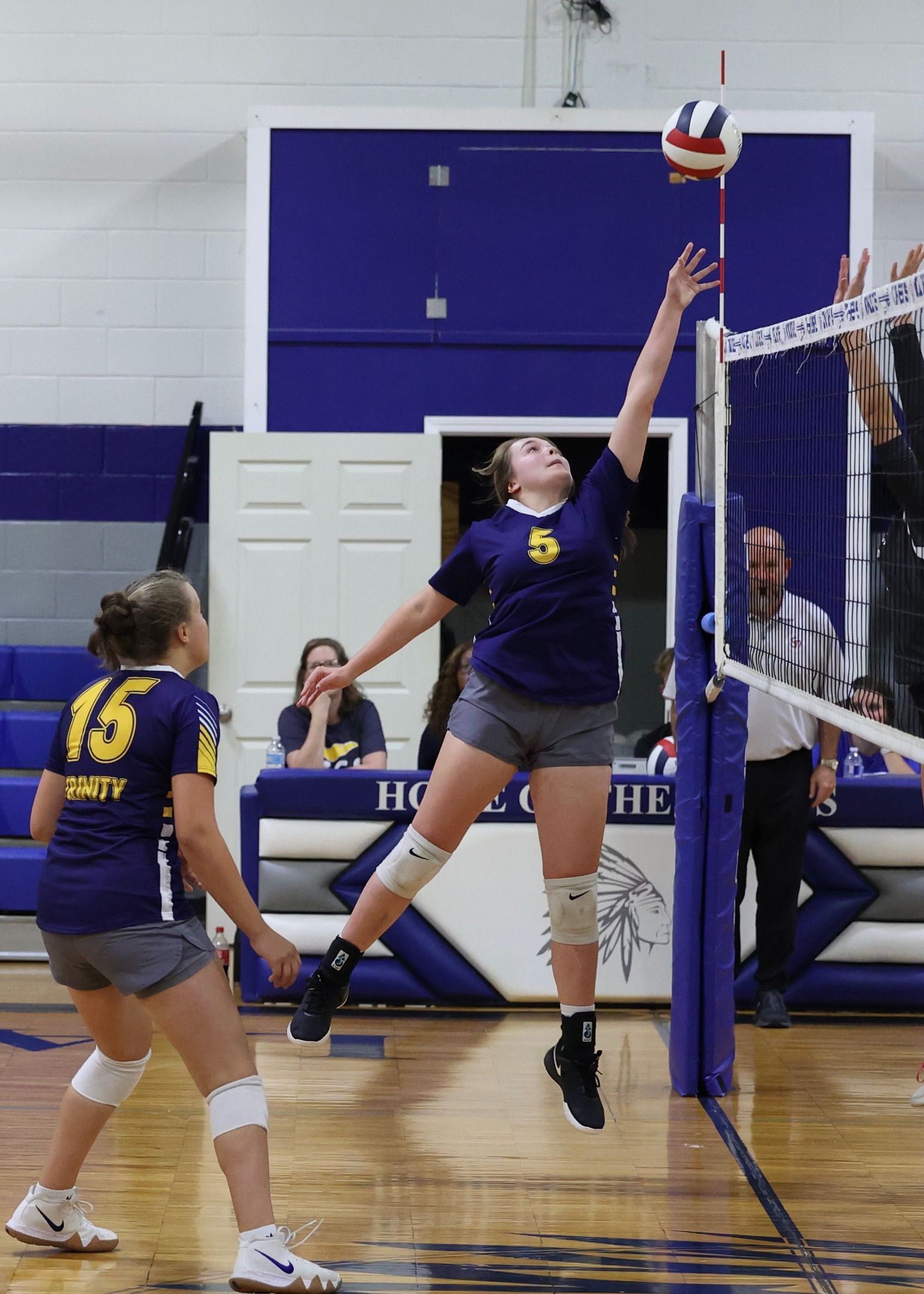 Two volleyball players jump at the net for a block and spike in a gymnasium.