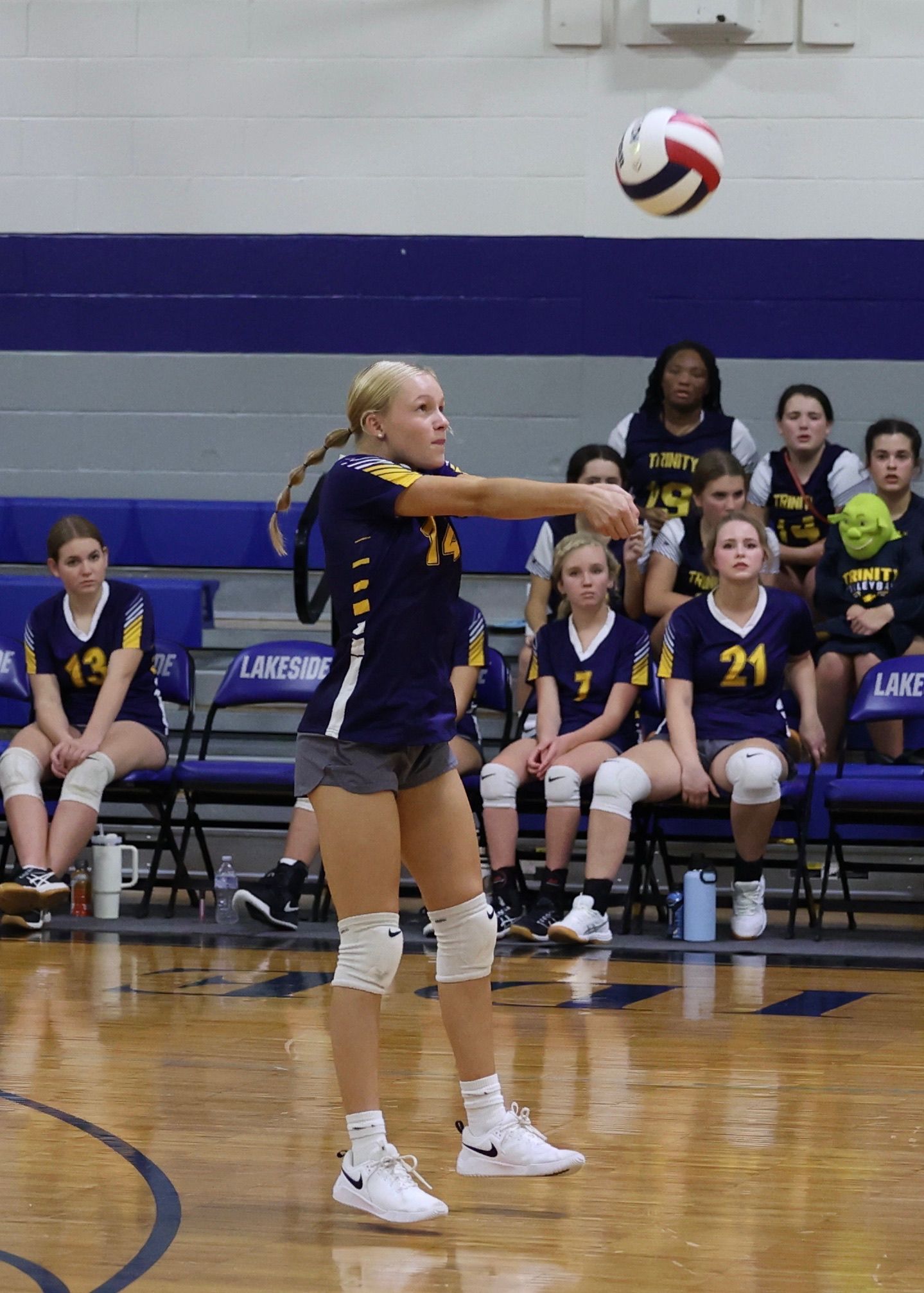 Volleyball player in navy uniform serving the ball on an indoor court, teammates seated behind her.