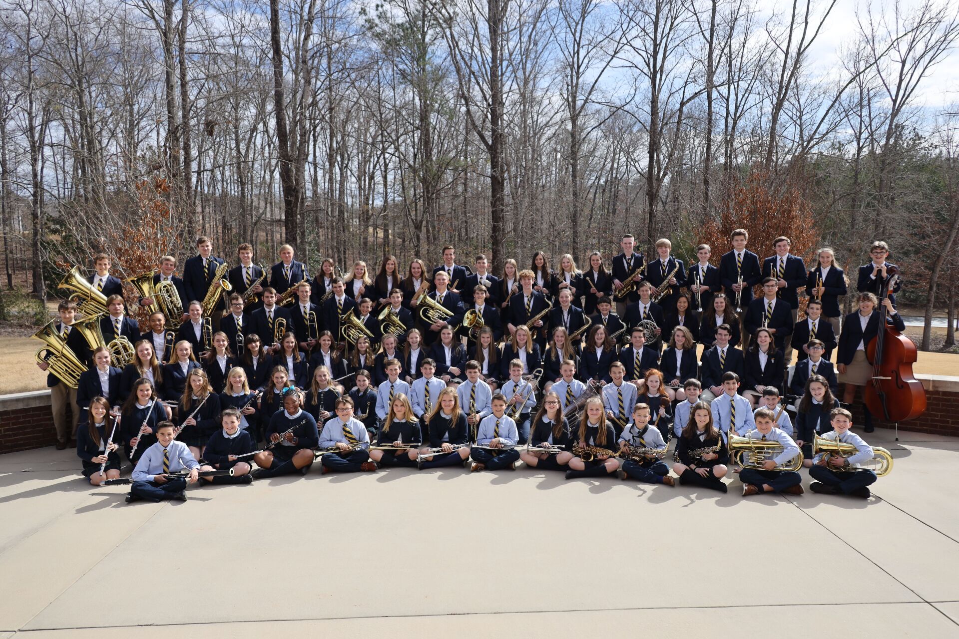 Large group portrait of an orchestra outdoors, holding instruments in front of leafless trees