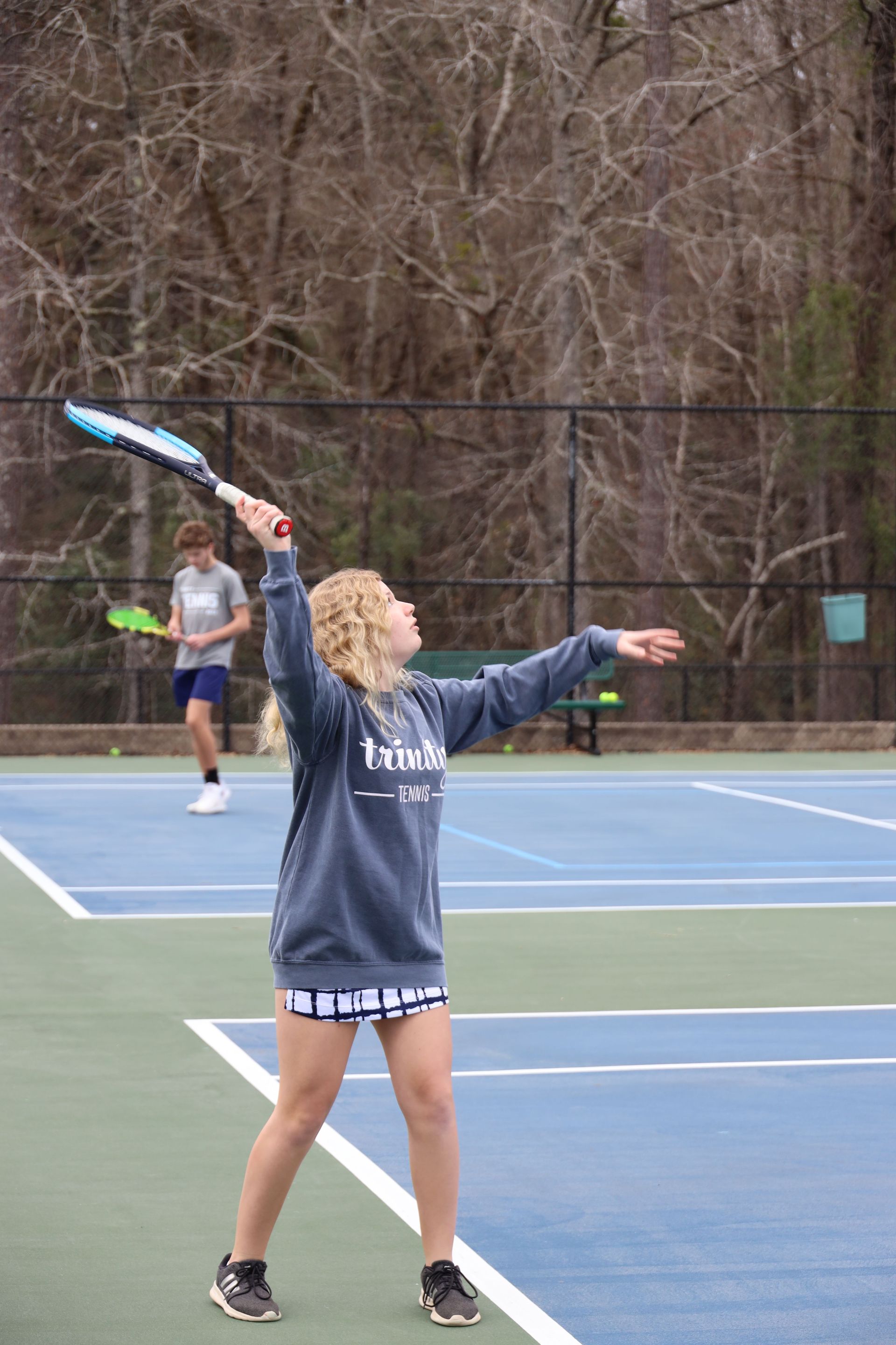 Tennis player in gray sweatshirt serves on an outdoor court, with another player in the background.