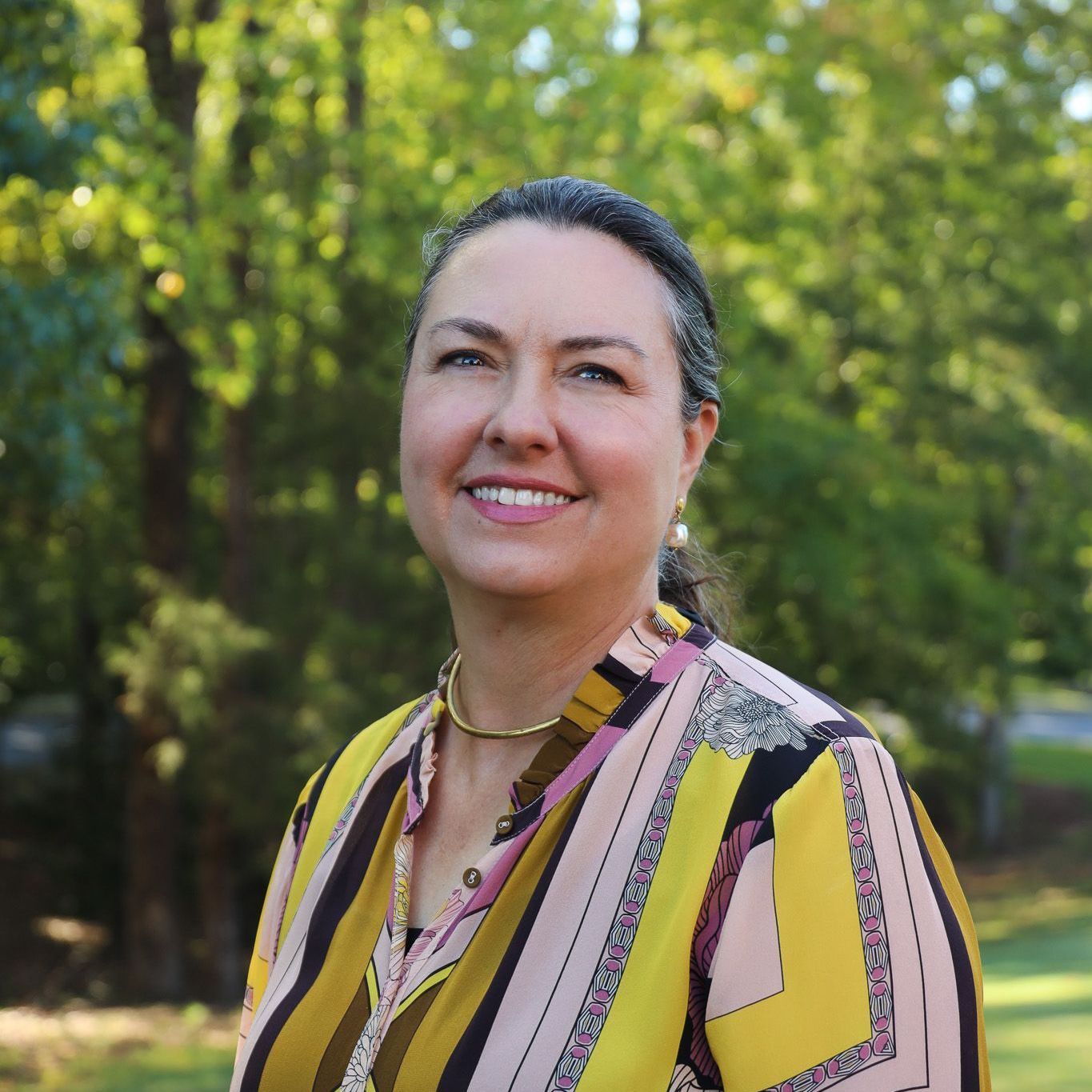 Smiling person in a yellow patterned shirt outdoors with green trees in the background