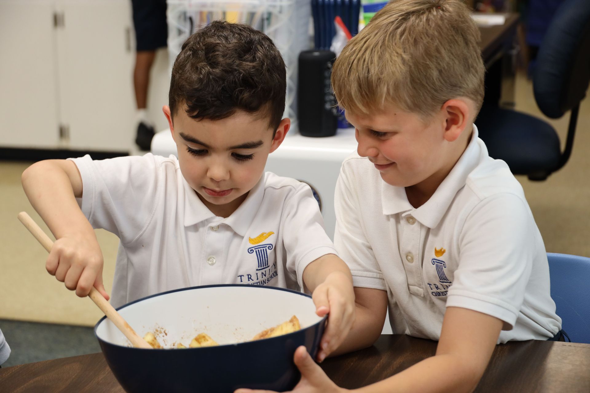 Two children in white shirts stirring a bowl together at a table in a classroom.