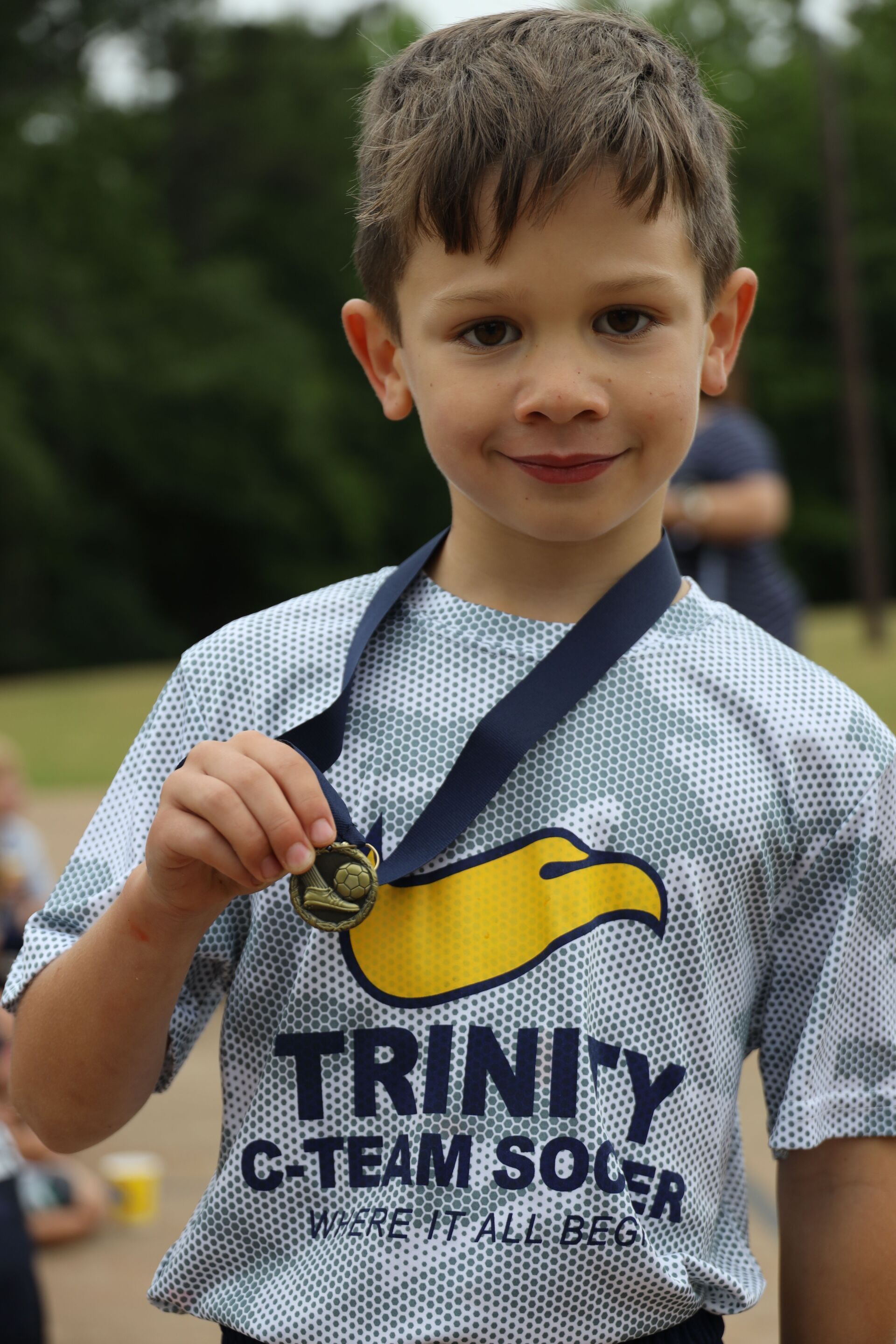 Child in a Trinity G-Team Soccer jersey holding up a medal outdoors, smiling at the camera.