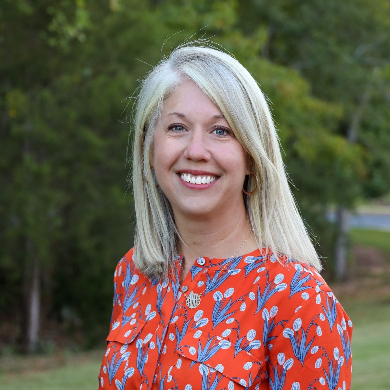 Smiling blonde woman in a red floral blouse standing outdoors with green trees behind her