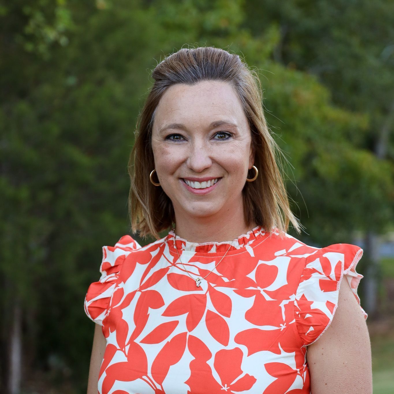 Smiling woman in a coral-and-white floral blouse standing outdoors against a blurred green background