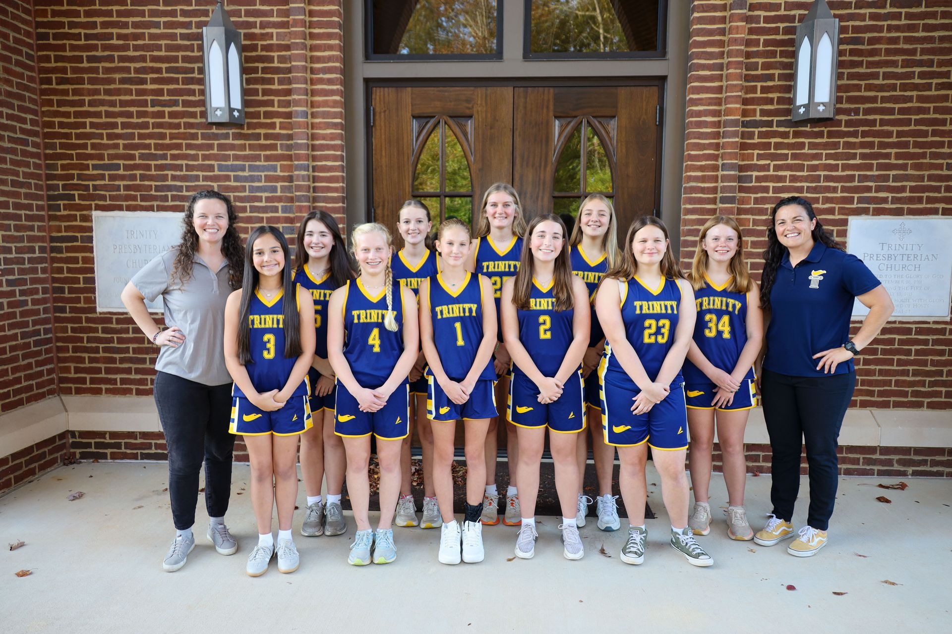 Basketball team posing in blue and yellow uniforms with two coaches in front of a brick building