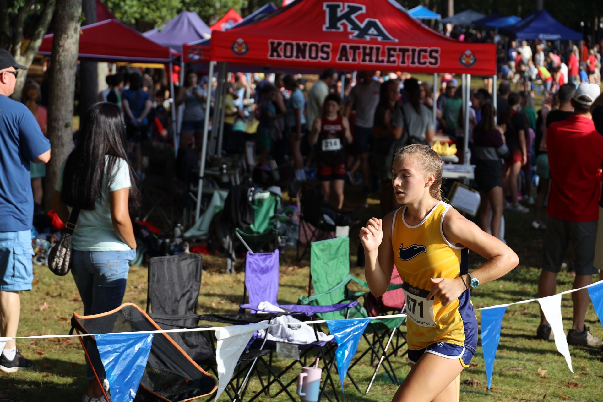 Runner in yellow racing past a Konos Athletics tent at a cross-country meet