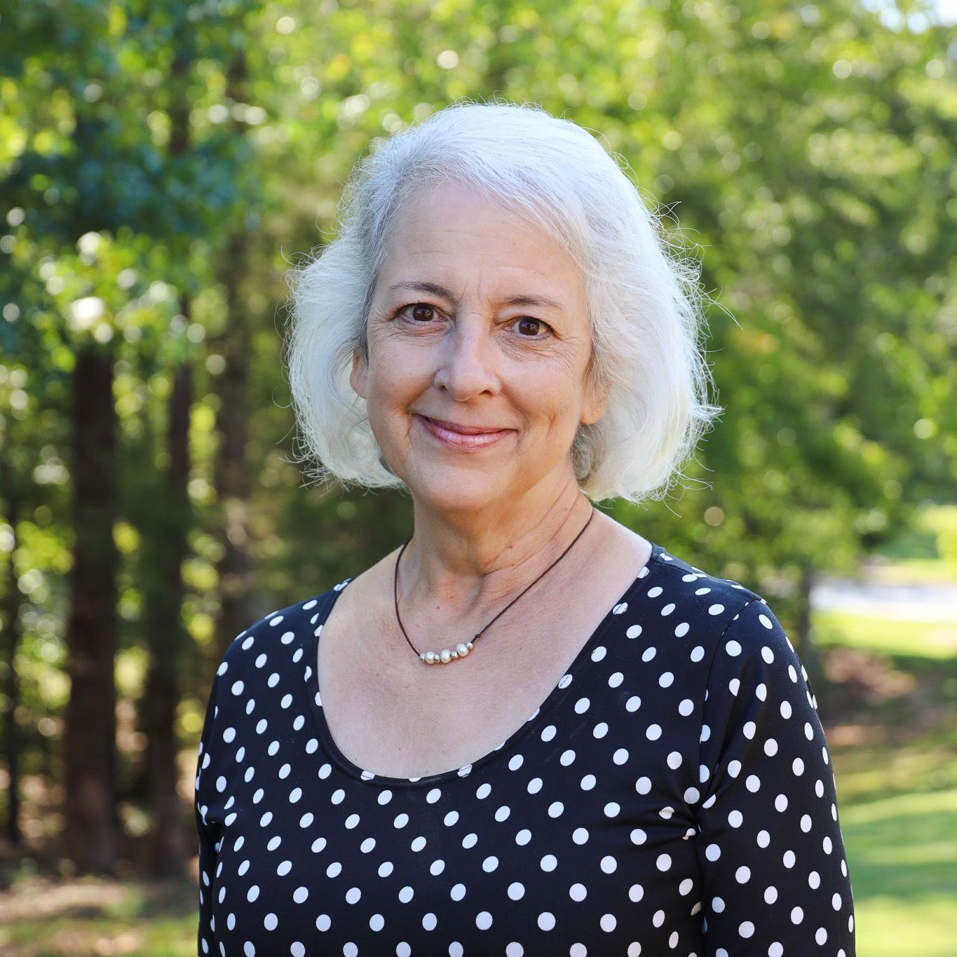 Smiling woman in a black polka dot top standing outdoors against a green, sunlit background