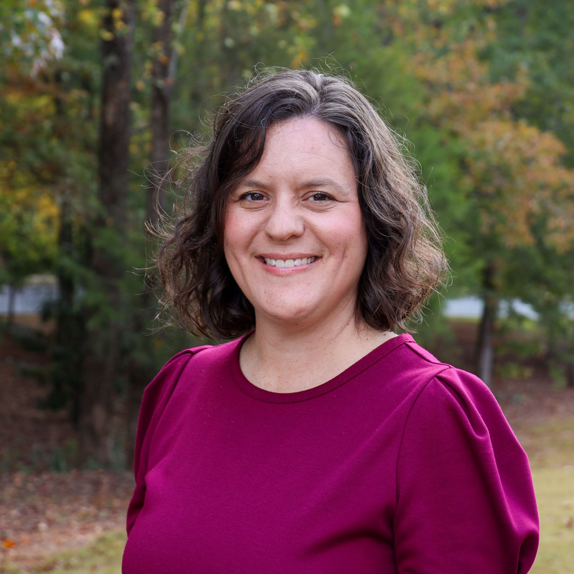 Smiling woman in a magenta top standing outdoors with trees in the background