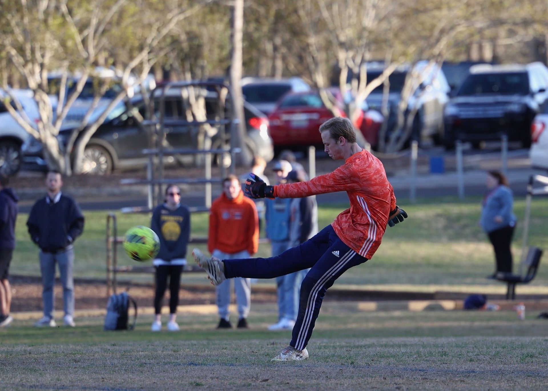 Person in red jacket kicking a ball on a field, with spectators and trees in the background.