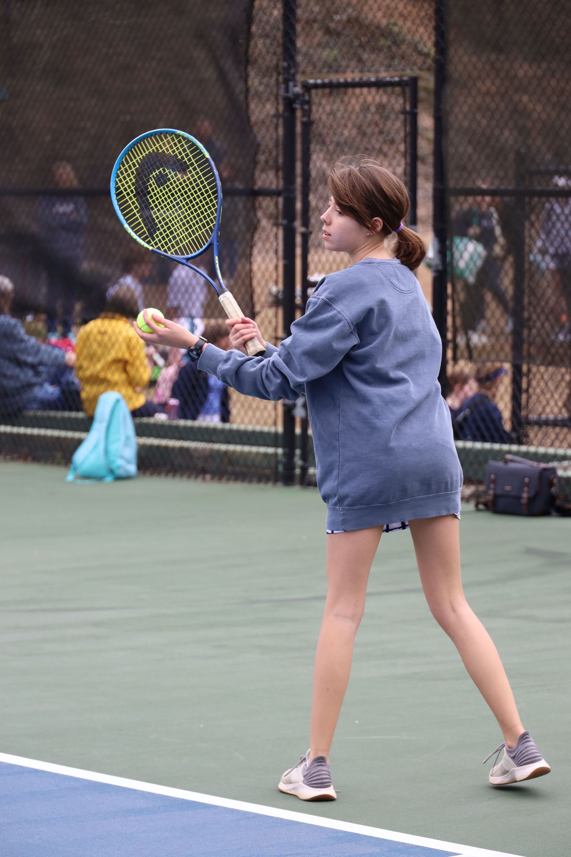 Person in blue jacket swings a tennis racket on an outdoor court.