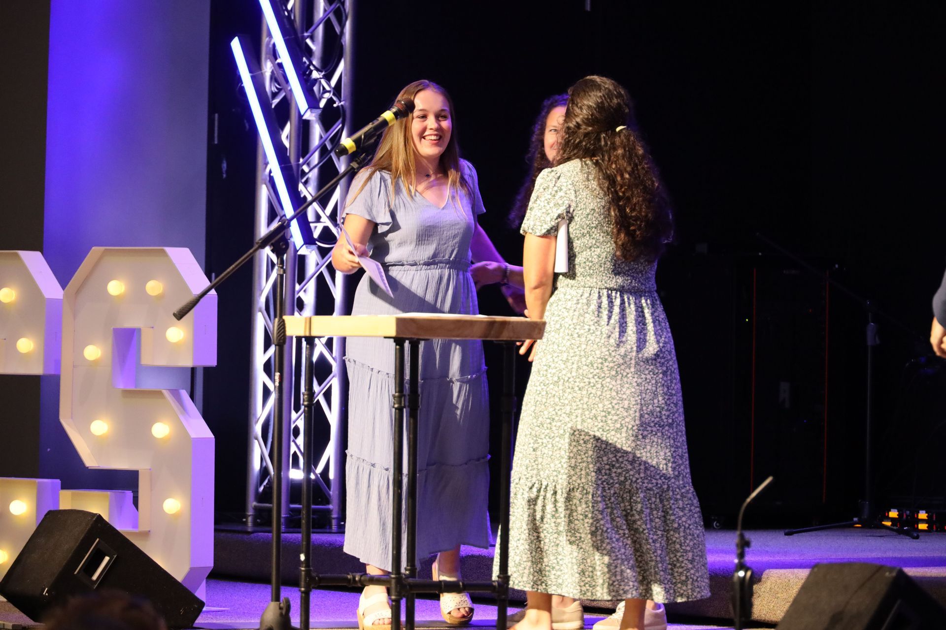 Two women in light dresses talking on a lit stage beside a large illuminated letter S