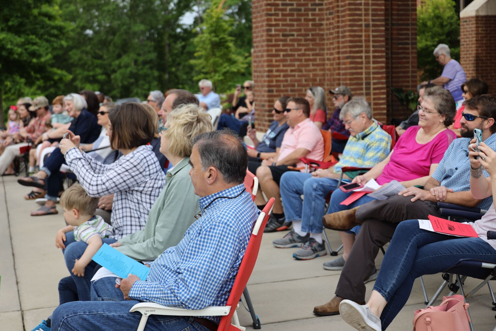 Audience seated outdoors at an event, with a brick building and trees in the background