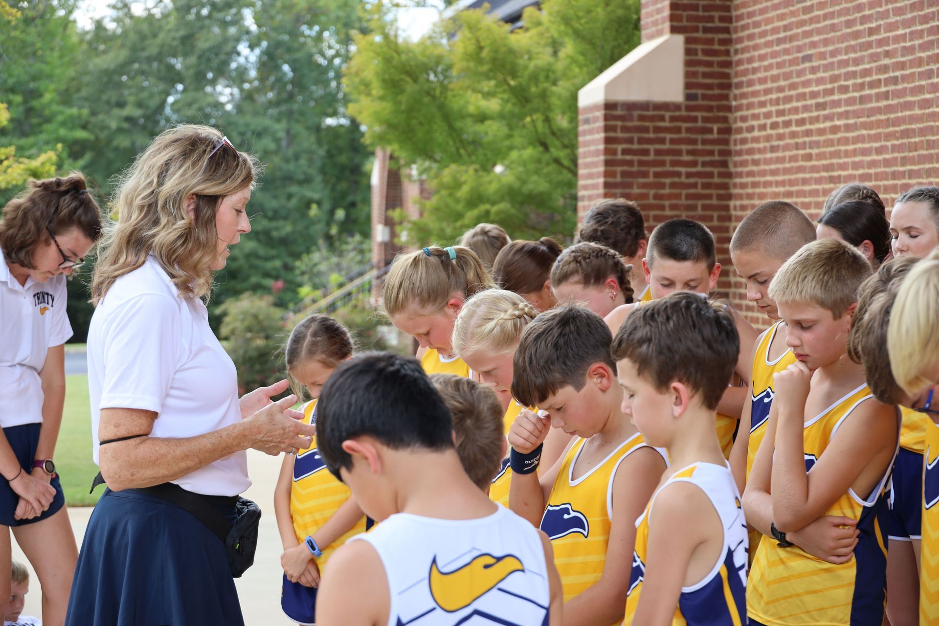 Students in yellow uniforms stand in line outdoors while an adult speaks to them near a brick building.