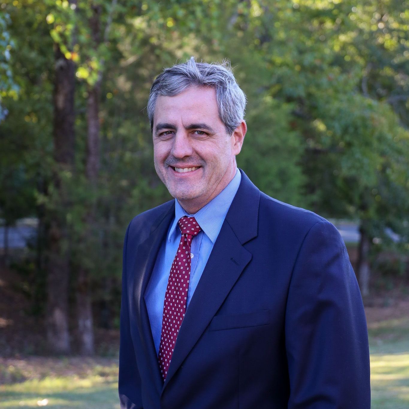 Man in a navy suit and red tie standing outdoors, smiling against a wooded background