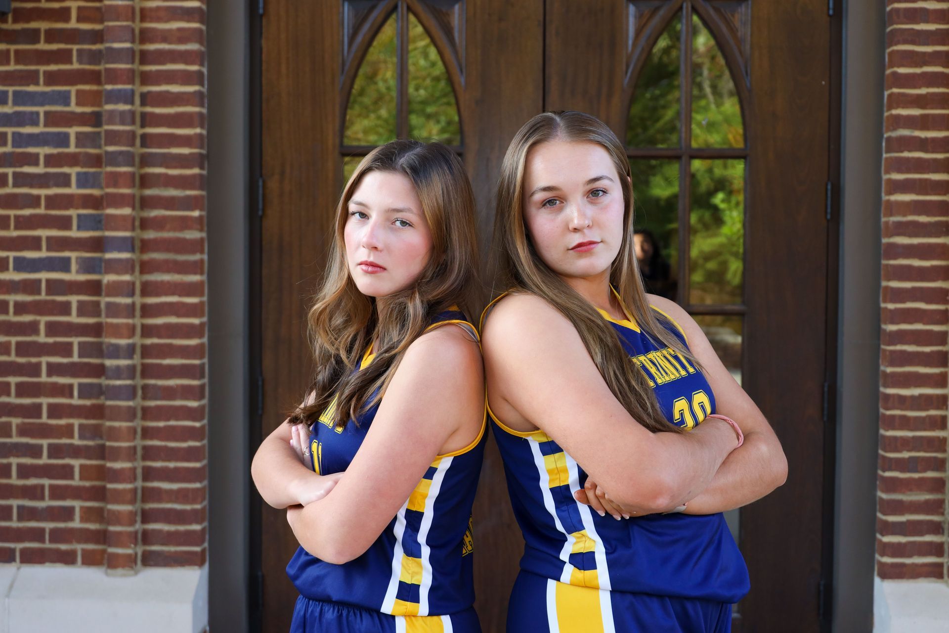 Two athletes in blue and yellow uniforms stand back-to-back with arms crossed in front of brick arches.