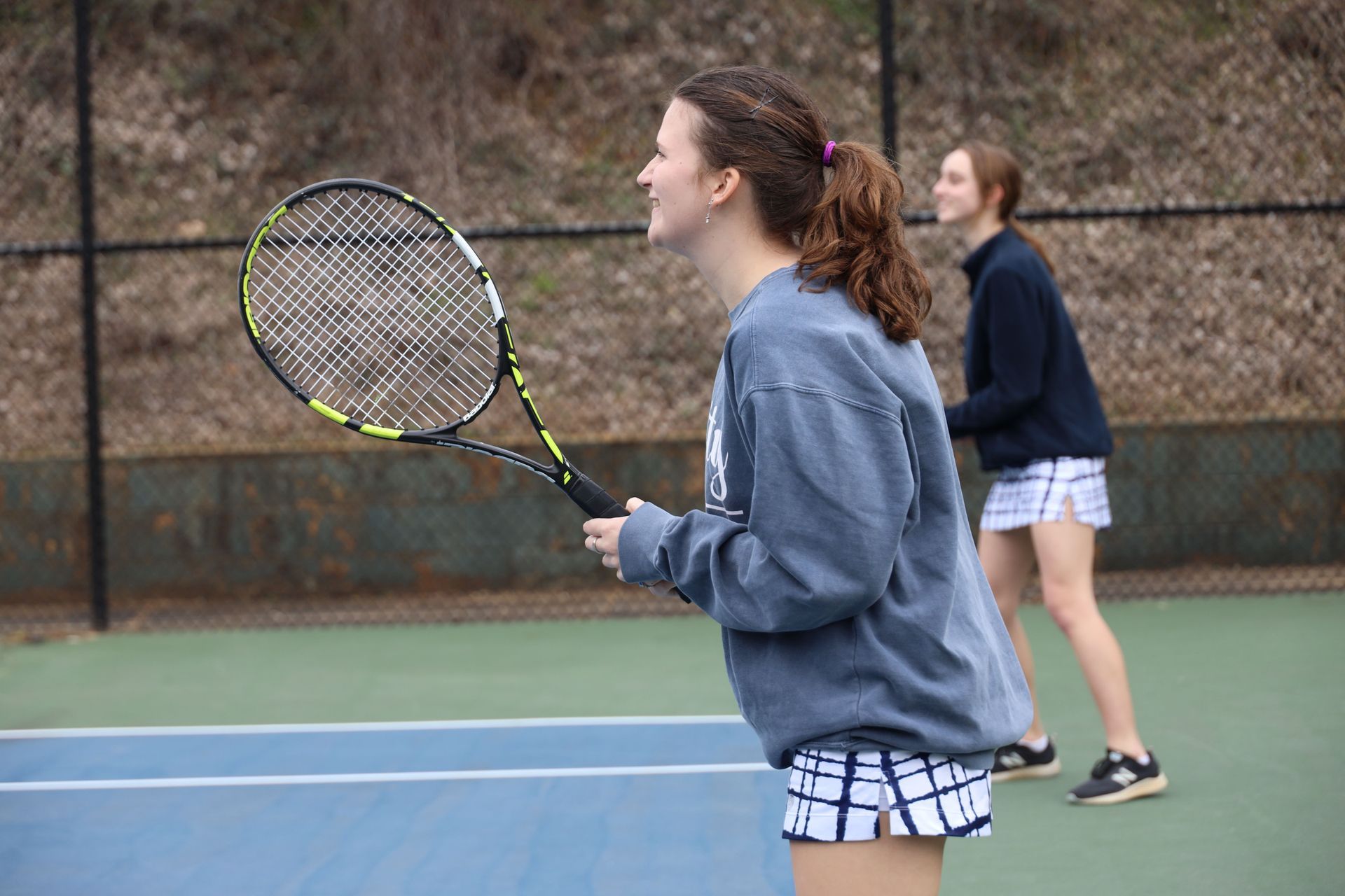 Two tennis players on an outdoor court, one in the foreground holding a racket ready to play.
