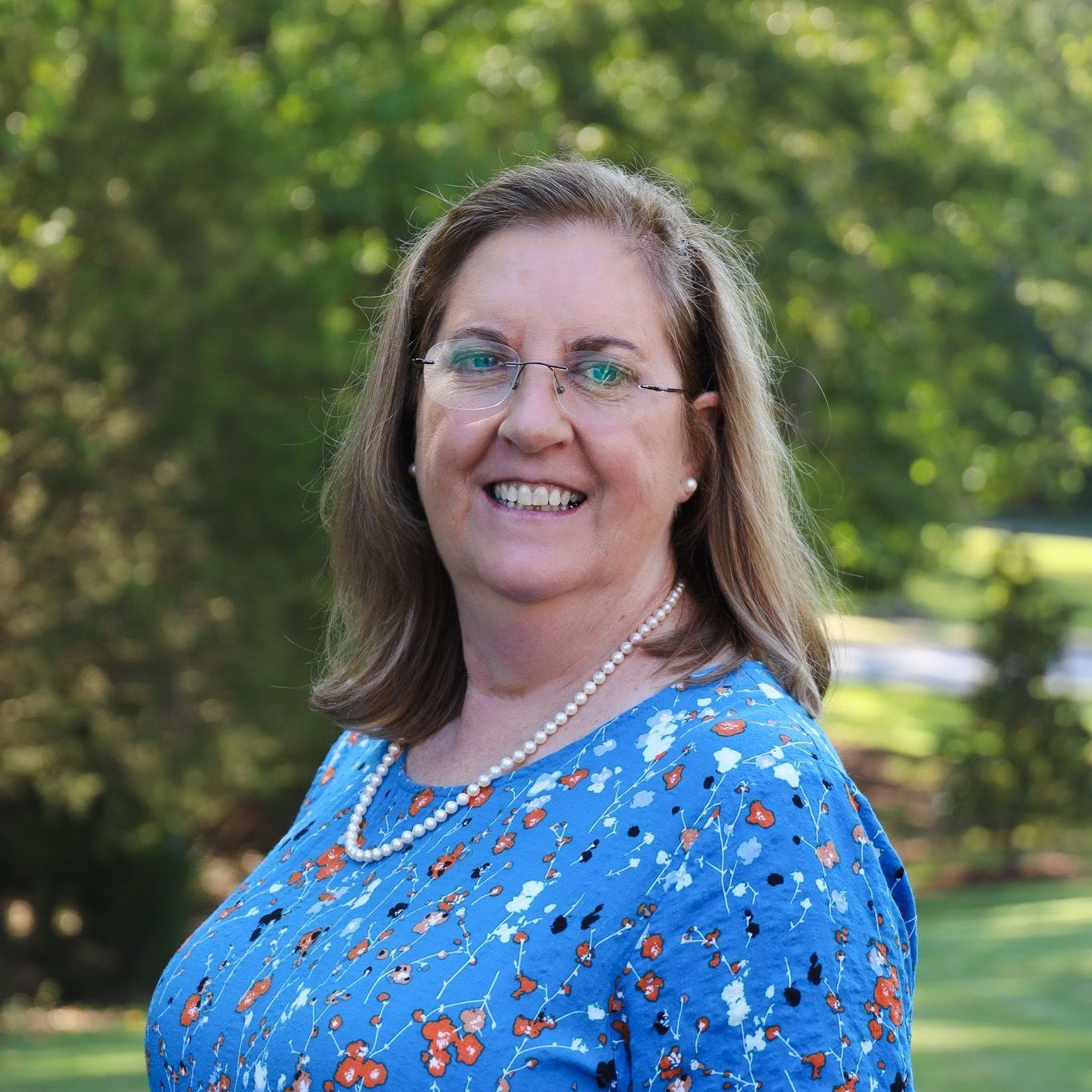 Smiling woman in blue floral top and pearl necklace outdoors with trees in the background