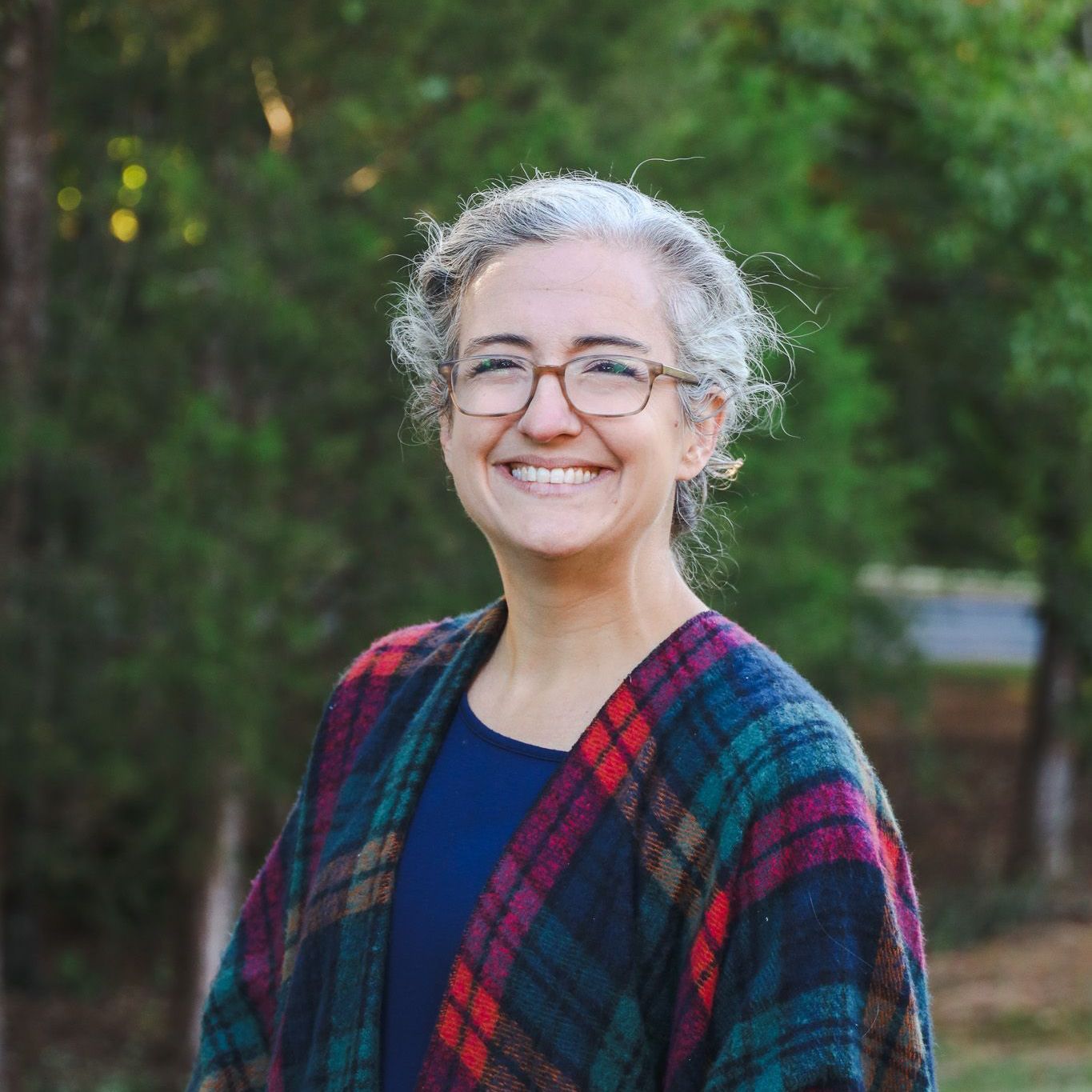 Smiling person with glasses wearing a red-and-blue plaid shawl outdoors against a green background