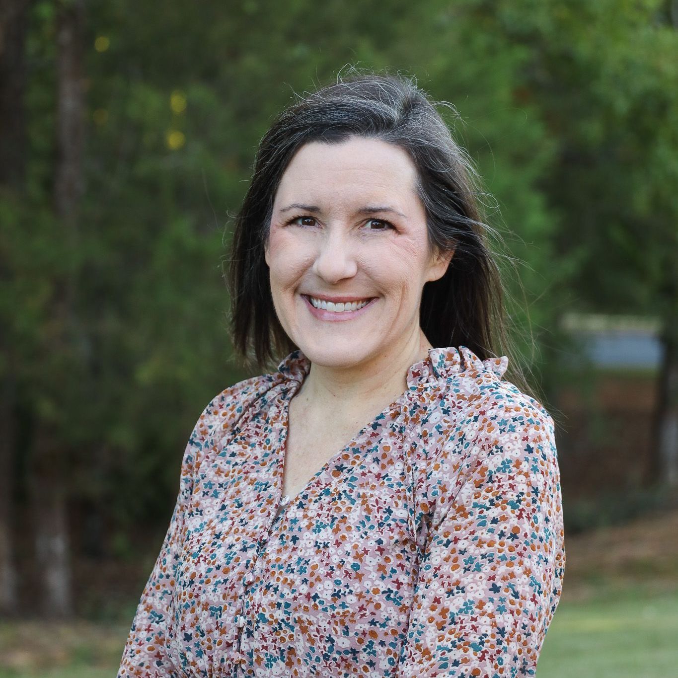 Smiling woman in a floral blouse outdoors with blurred green trees in the background