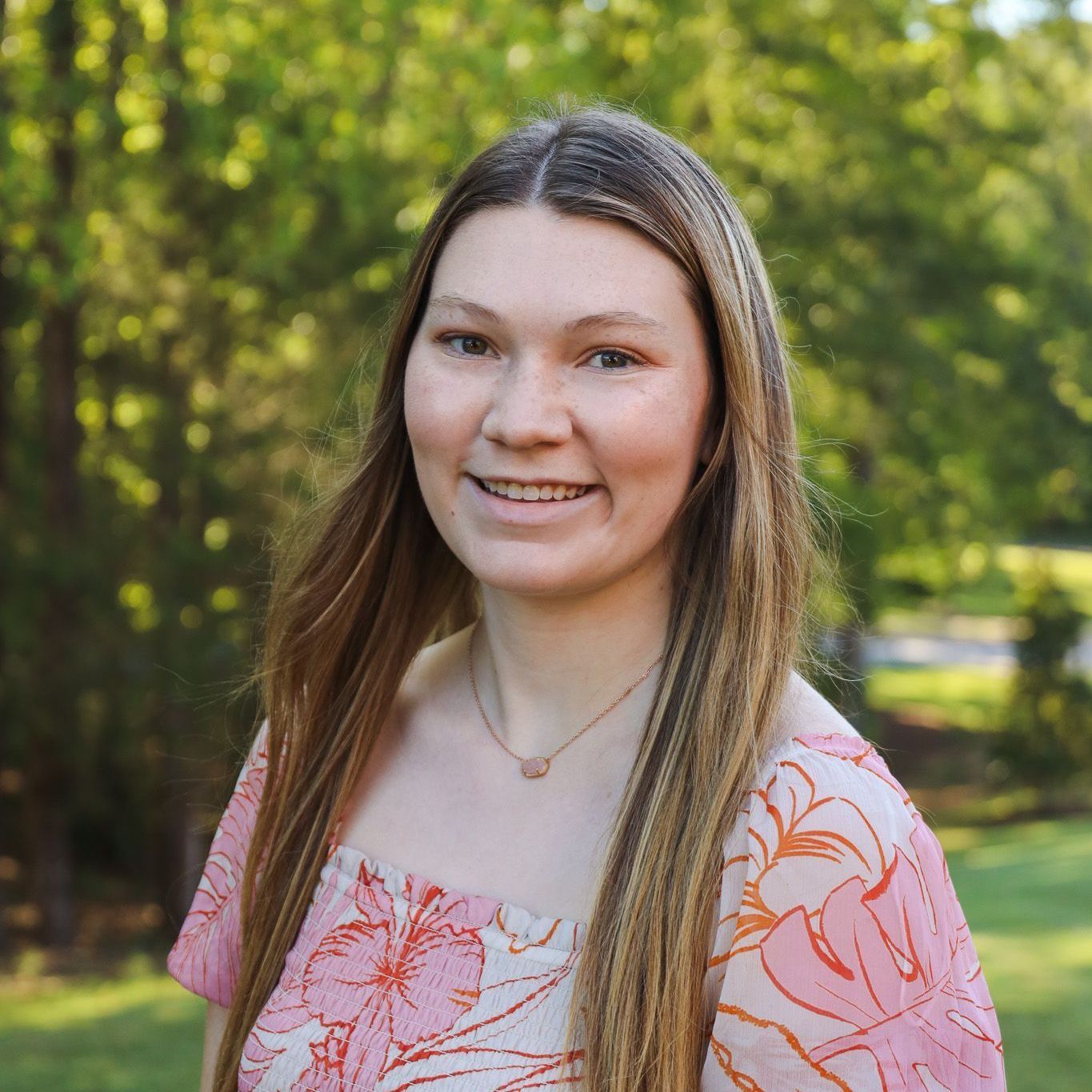 Smiling person outdoors in a pink floral top, standing in front of green trees and sunlight