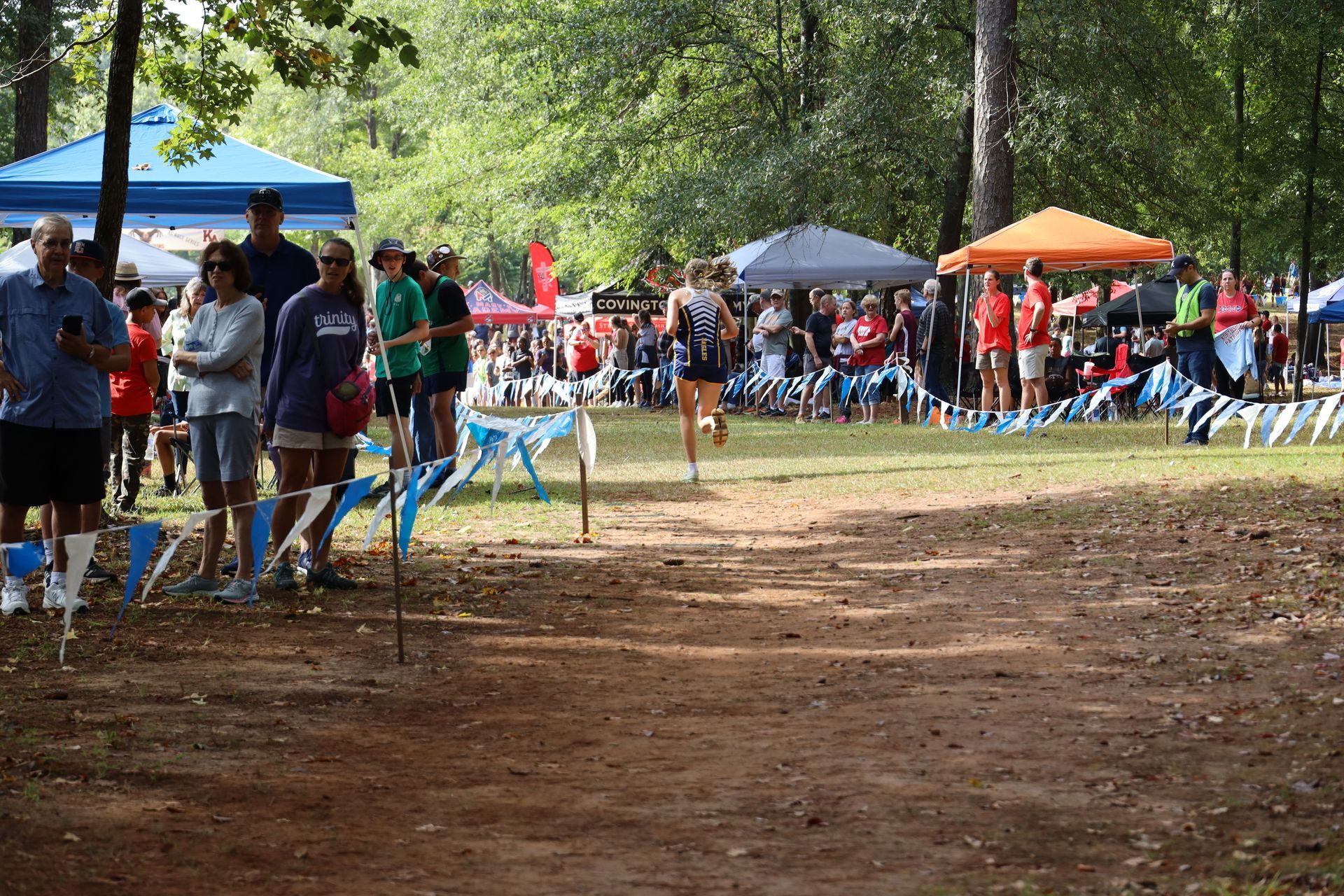 People gathered at an outdoor event near a dirt path, with colorful tents and bunting in the background.