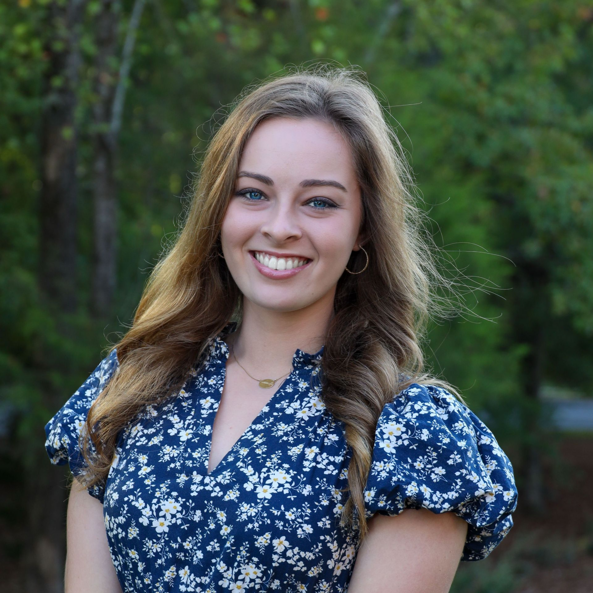 Smiling woman in a blue floral dress standing outdoors with greenery behind her