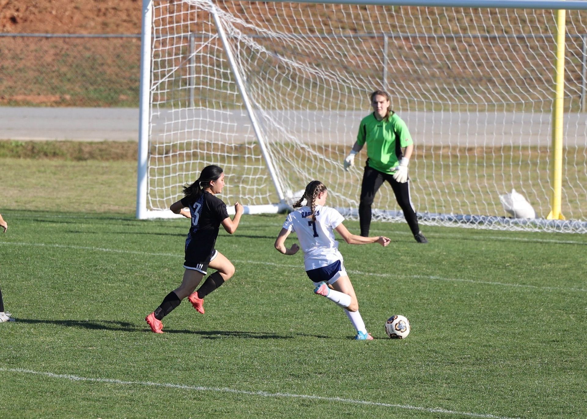 Two soccer players chasing the ball near goal, with a goalkeeper ready in the net