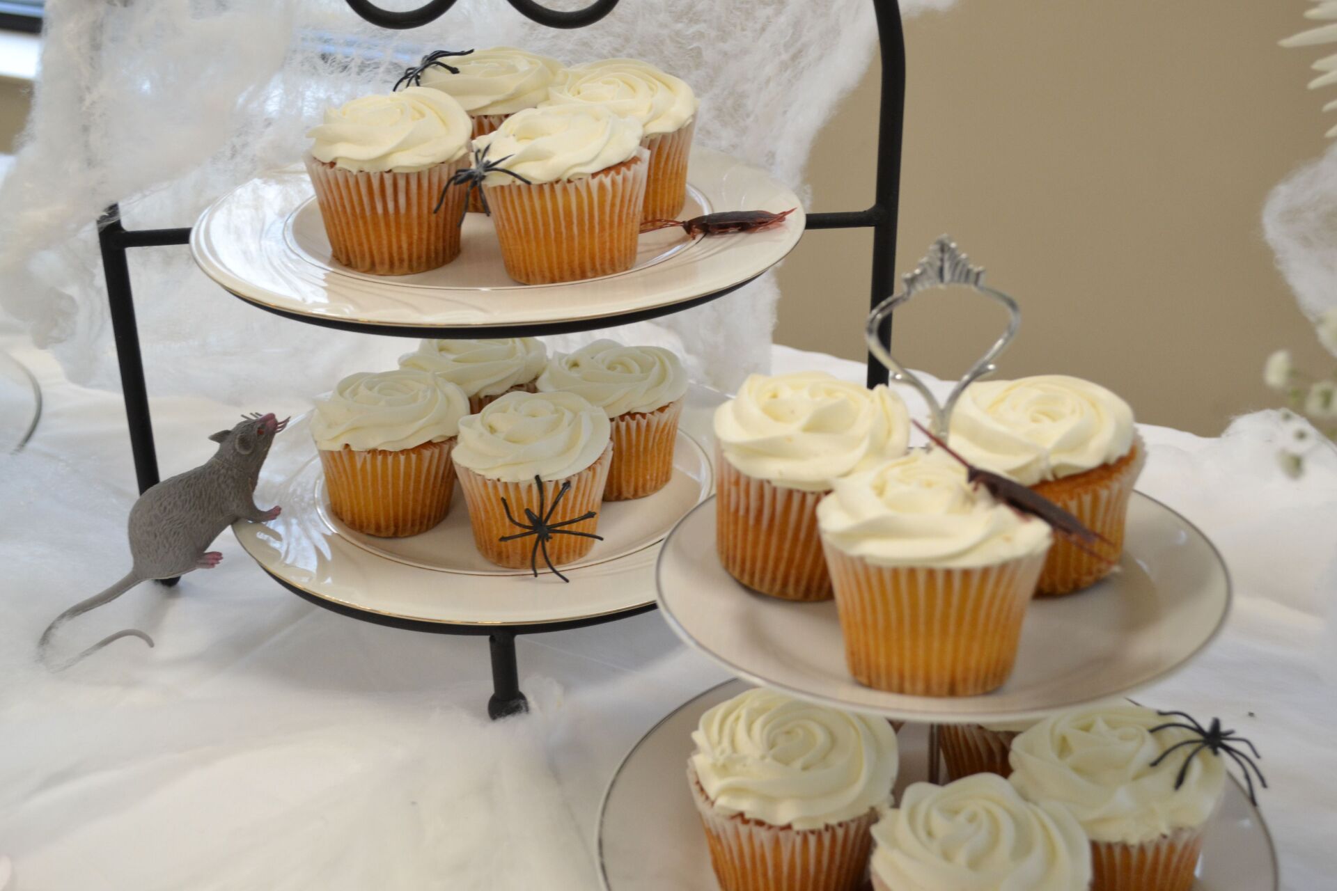 Tiered dessert stand with white-iced cupcakes and small black spiders on a white tablecloth