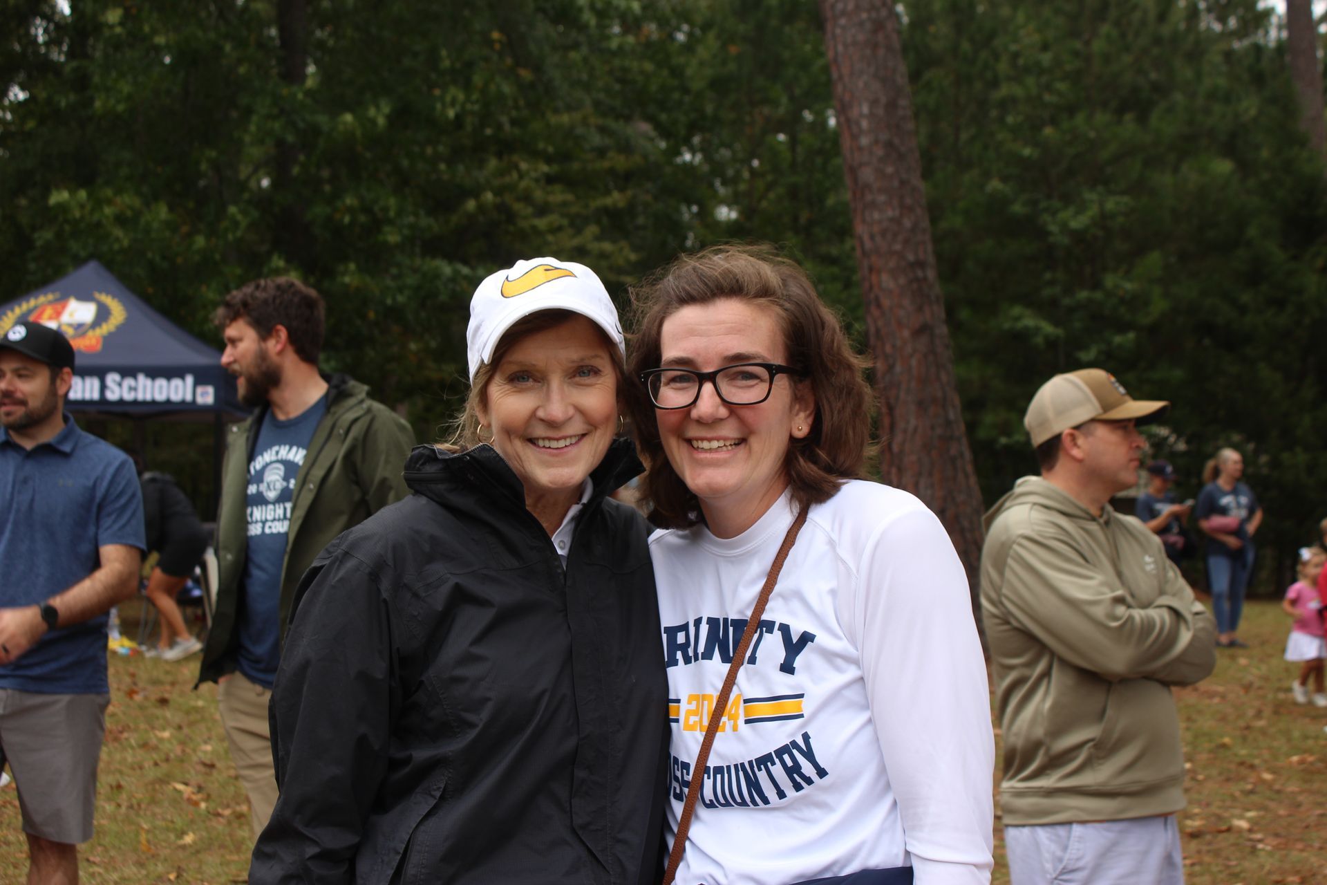 Two women smiling outdoors, one in a black jacket and cap, the other in a white T-shirt and glasses.