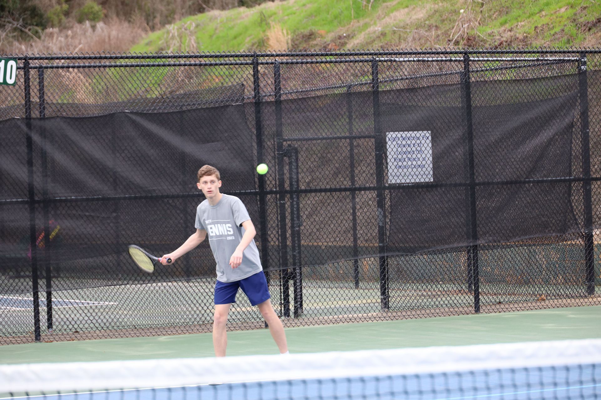 Tennis player in gray shirt and blue shorts prepares to hit a ball on an outdoor court.