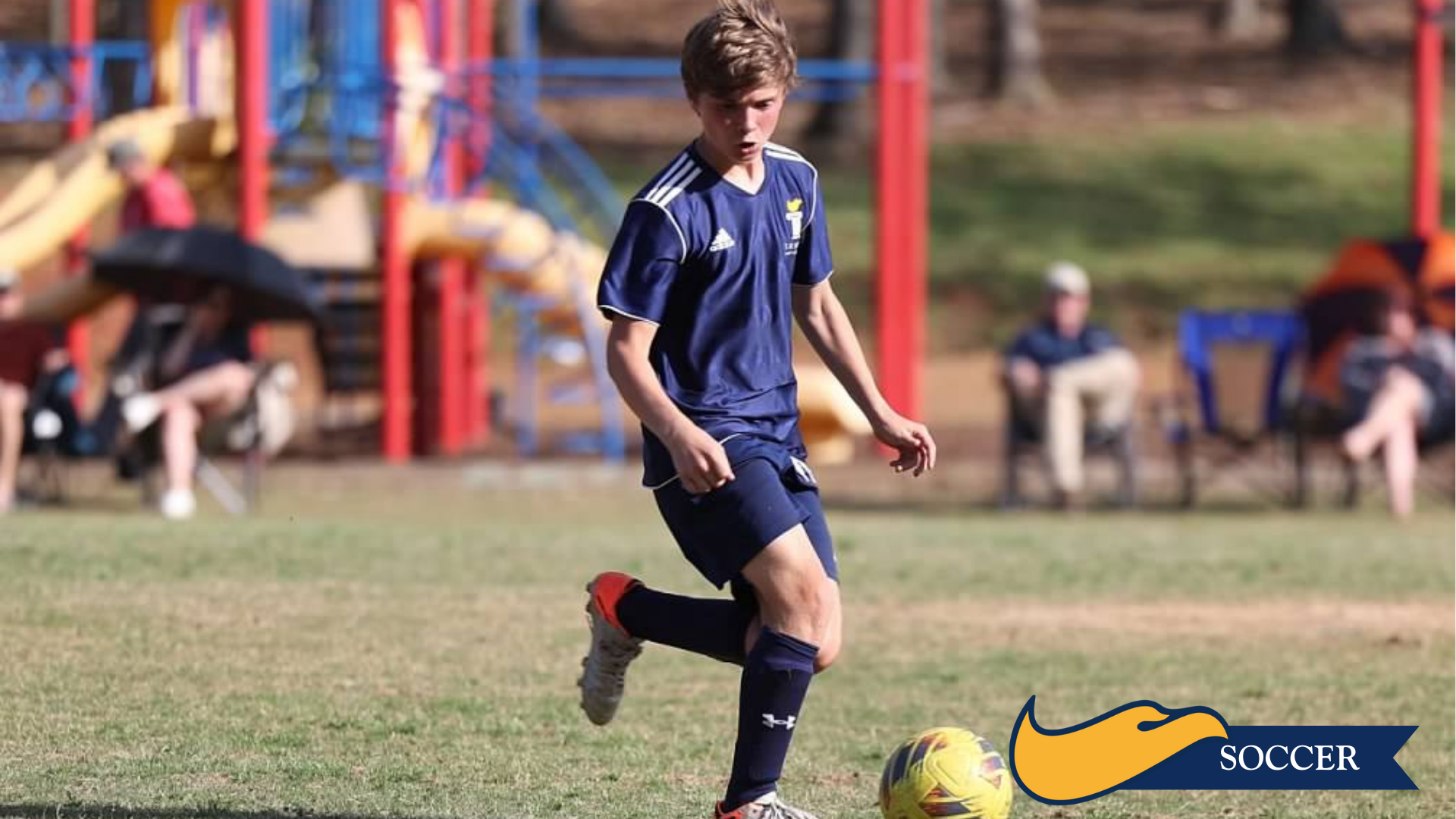 Soccer player in navy uniform dribbling a yellow ball on a field with spectators in the background