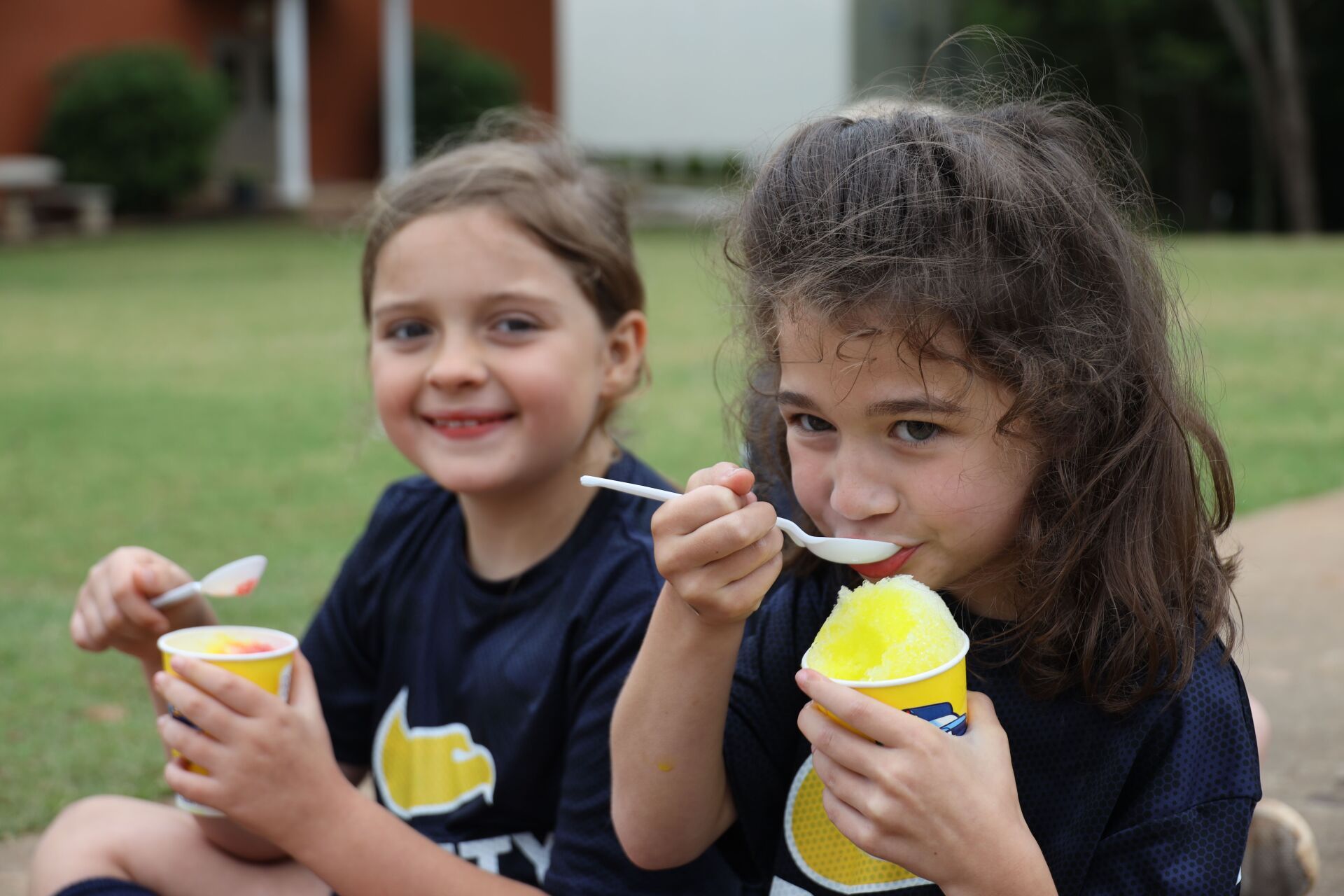 Two children in navy shirts eating yellow ice cream outdoors on a lawn